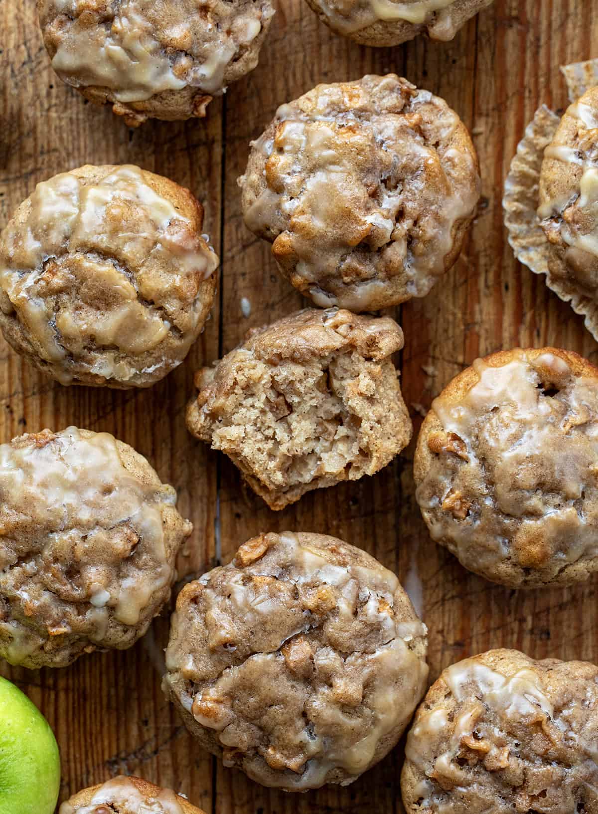 Old Fashioned Apple Muffins are moist, dense muffins loaded with apples and drizzled with a maple glaze, perfect for breakfast, brunch, or as a delightful dessert option. Enjoy the comforting flavors of fall and the sweetness of maple in every delicious bite! Apples Muffins on a Wooden Cutting Board with One Muffin Halved to Show Inside from Overhead.