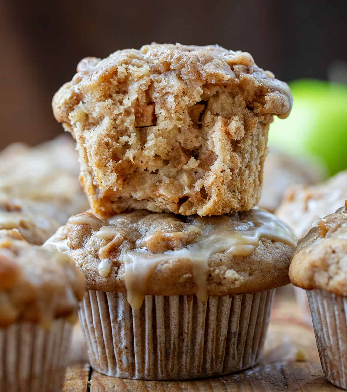 Old Fashioned Apple Muffins are moist, dense muffins loaded with apples and drizzled with a maple glaze, perfect for breakfast, brunch, or as a delightful dessert option. Enjoy the comforting flavors of fall and the sweetness of maple in every delicious bite! Half Apple Muffin on Top of Another Muffin on a Cutting Board with an Apple in the Back.