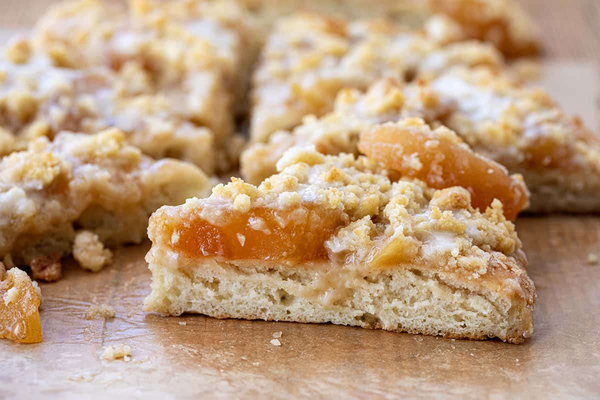 Apple Dessert Pizza is a delightful and unique treat that combines the flavors of a classic apple pie with the fun and convenience of a pizza. It starts with a soft focaccia bread crust topped with cinnamon butter spread, apple pie filling, and crumble topping for a little crunch. And then, a simple glaze is drizzled over the top. It will satisfy your sweet tooth with its cinnamon apple flavor and sweet glaze! Close up of a Slice of Apple Dessert Pizza.