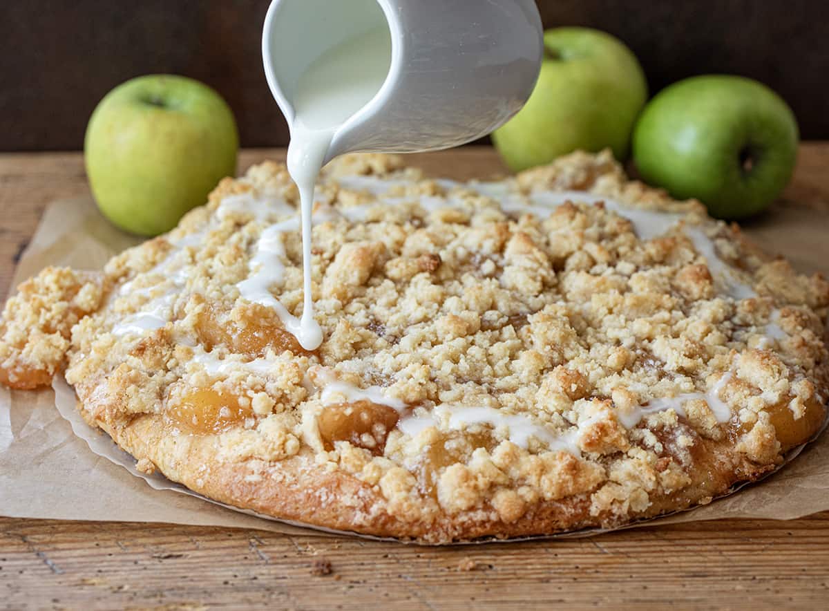 Apple Dessert Pizza is a delightful and unique treat that combines the flavors of a classic apple pie with the fun and convenience of a pizza. It starts with a soft focaccia bread crust topped with cinnamon butter spread, apple pie filling, and crumble topping for a little crunch. And then, a simple glaze is drizzled over the top. It will satisfy your sweet tooth with its cinnamon apple flavor and sweet glaze! Pouring Glaze Over Apple Dessert Pizza.