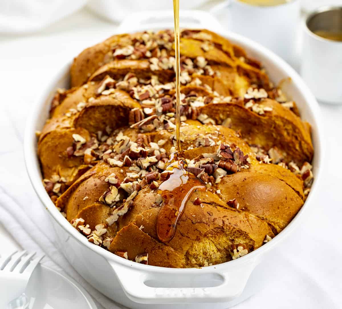 Pumpkin French Toast Casserole is slices of Challah bread dipped into a pumpkin-flavored custard mixture, sprinkled with pecans, and baked for a warm breakfast or brunch. It's a perfect recipe to prepare the night before and get in the oven the next morning! Adding Syrup to Pumpkin French Toast Casserole in White Pan on White Counter.