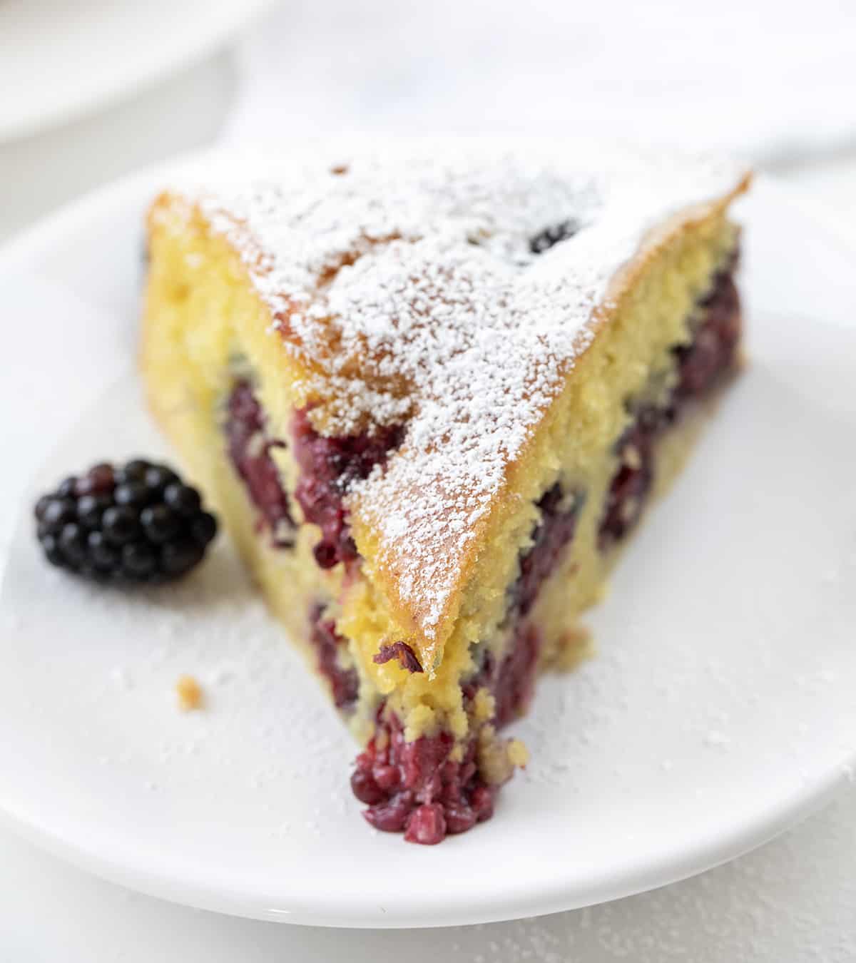 Blackberry Breakfast Cake is a cake filled with fresh blackberries, baked and then drenched in a warm sweet milk mixture for an incredibly moist and flavorful bite! It's a delicious morning treat alongside a cup of coffee, but regardless of the name, it's not just for breakfast! It can also be enjoyed as an afternoon snack or as a dessert after any meal! Piece of Blackberry Breakfast Cake on a White Plate with Fork.