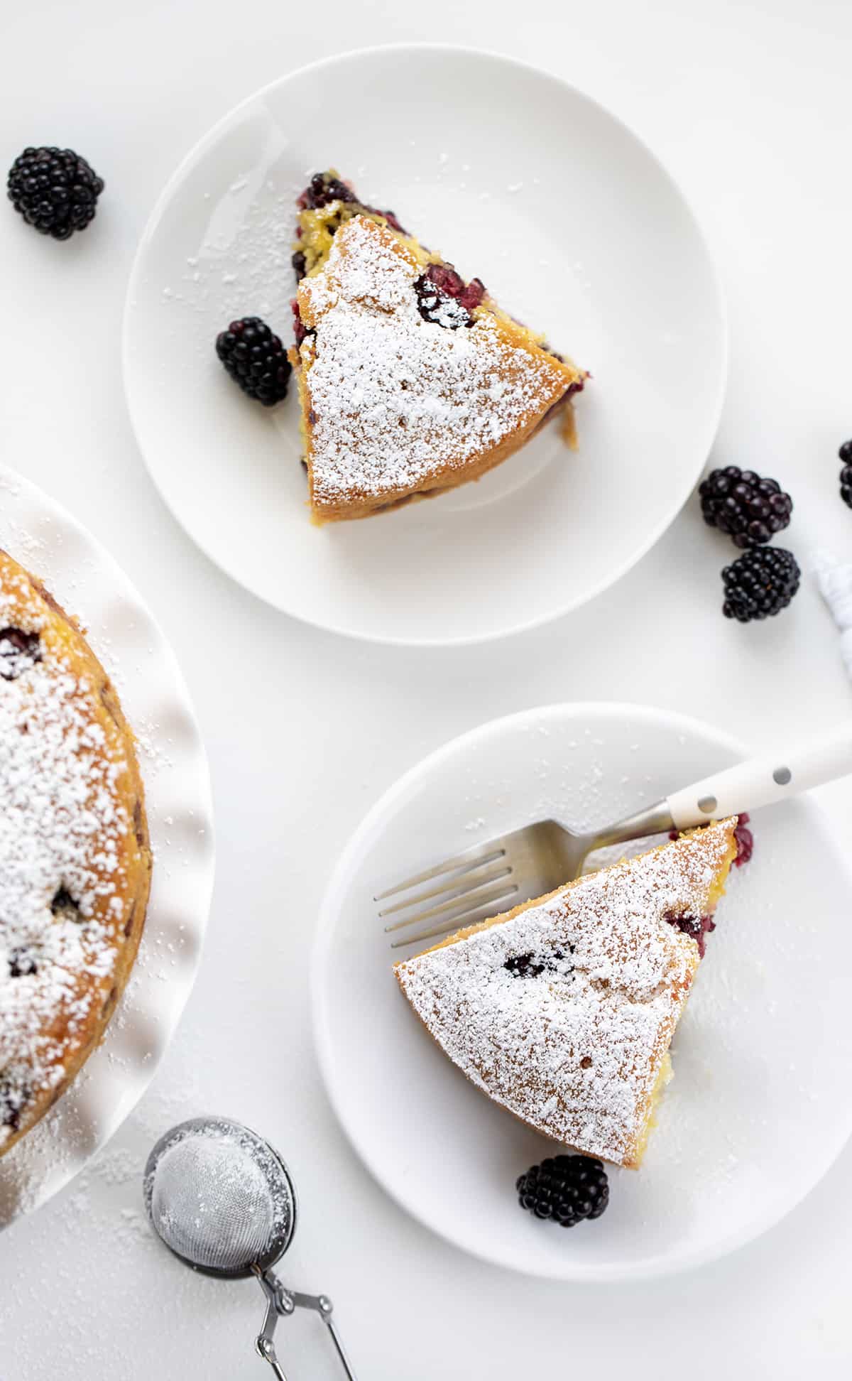 Blackberry Breakfast Cake is a cake filled with fresh blackberries, baked and then drenched in a warm sweet milk mixture for an incredibly moist and flavorful bite! It's a delicious morning treat alongside a cup of coffee, but regardless of the name, it's not just for breakfast! It can also be enjoyed as an afternoon snack or as a dessert after any meal! White Plates with Blackberry Breakfast Cake on Them with White Form and Fresh Blackberries on a White Counter.