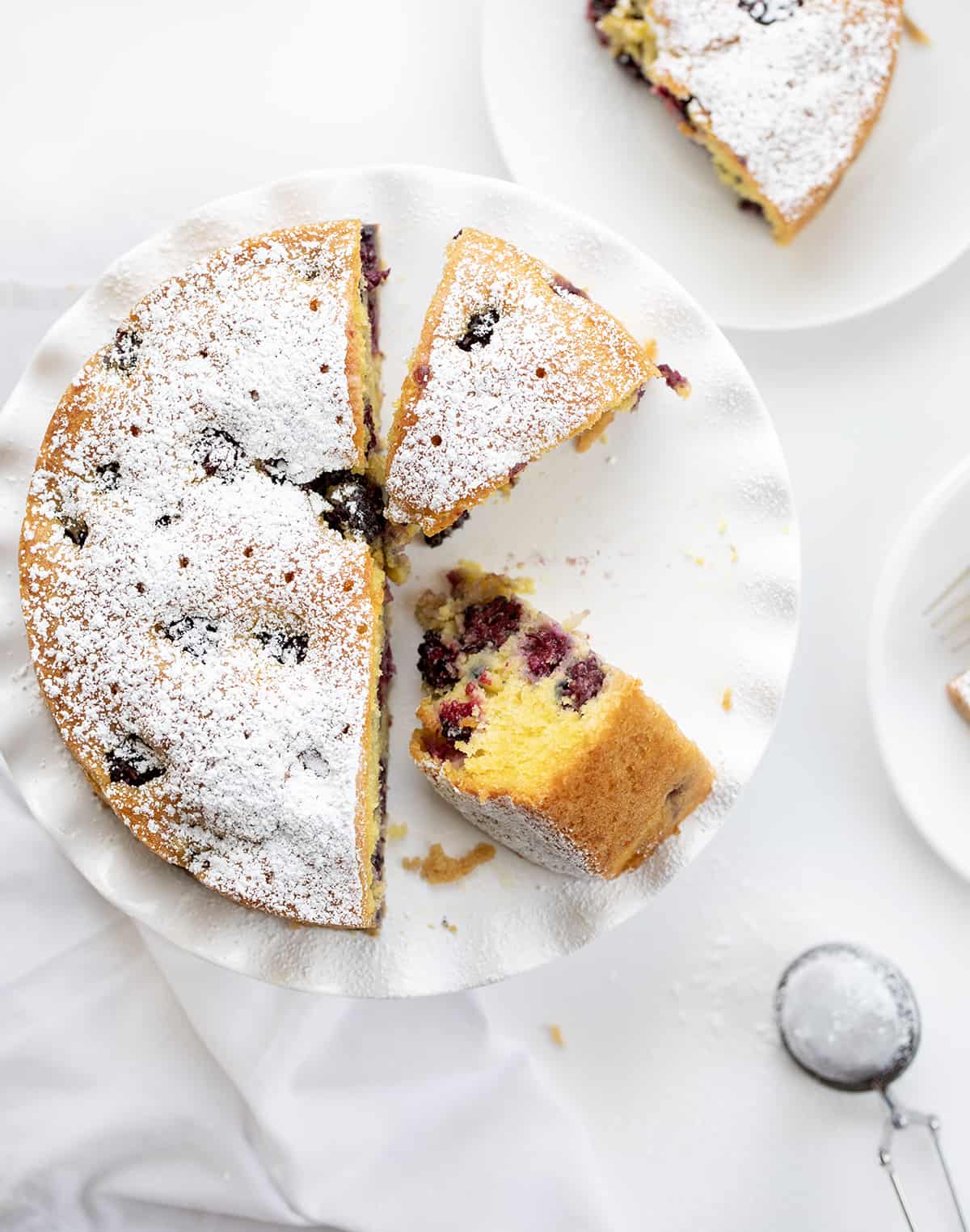 Blackberry Breakfast Cake is a cake filled with fresh blackberries, baked and then drenched in a warm sweet milk mixture for an incredibly moist and flavorful bite! It's a delicious morning treat alongside a cup of coffee, but regardless of the name, it's not just for breakfast! It can also be enjoyed as an afternoon snack or as a dessert after any meal! Blackberry Breakfast Cake on a Cake Stand with Some Pieces Removed and One Piece on its Side and Other plates Around.