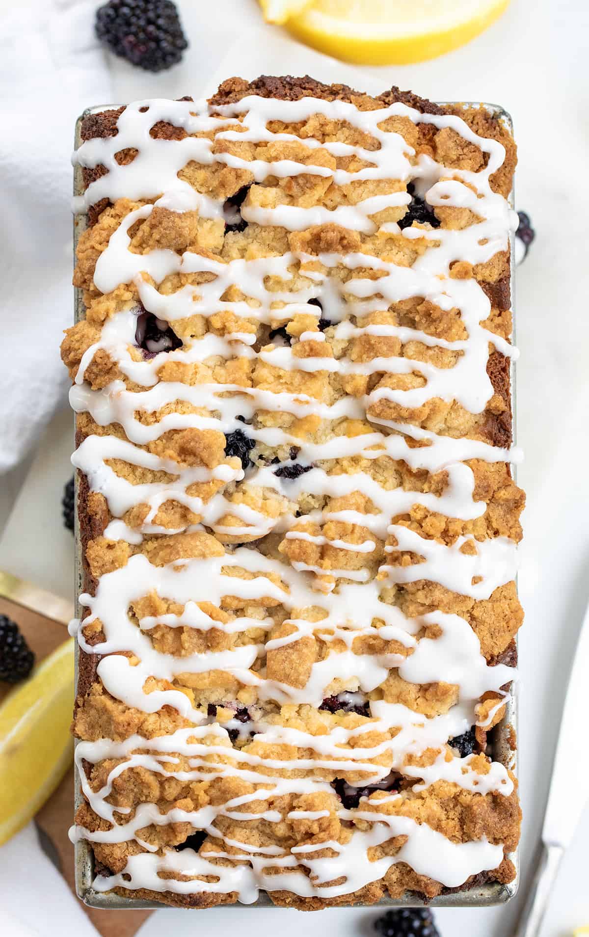 Blackberry Lemon Loaf is a moist, cake-like loaf with lots of fresh blackberries and tangy lemon flavor, topped with a buttery, crumbly topping. And then, a sweet glaze is drizzled over the loaf. Enjoy a slice anytime you crave a sweet and zesty treat Blackberry Lemon Loaf on a White Counter with Glaze.