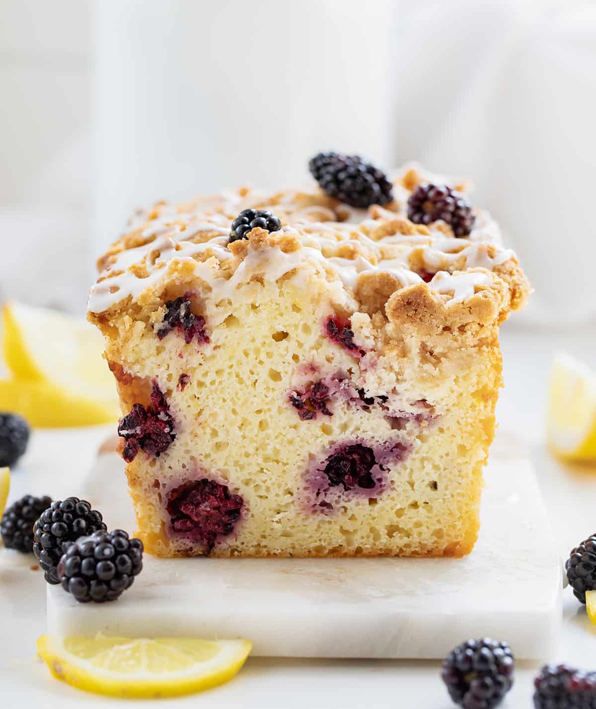 Blackberry Lemon Loaf is a moist, cake-like loaf with lots of fresh blackberries and tangy lemon flavor, topped with a buttery, crumbly topping. And then, a sweet glaze is drizzled over the loaf. Enjoy a slice anytime you crave a sweet and zesty treat Blackberry Lemon Loaf on a Marble Cutting Board Surrounded by Blackberries and Lemon.