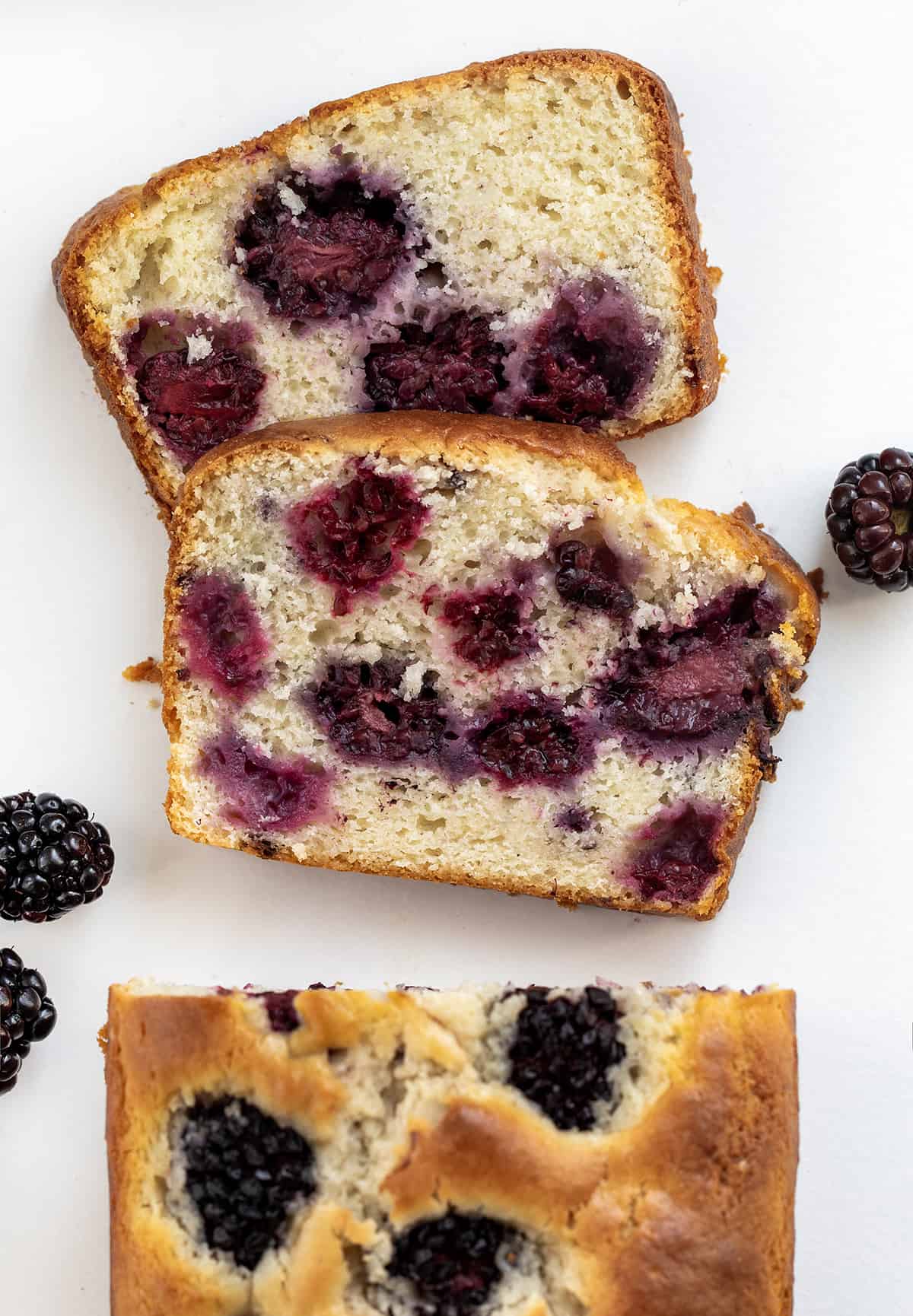 Blackberry Loaf is a soft and tender quick bread loaded with fresh blackberries and drizzled with an optional sweet glaze. Whether you're planning a casual brunch, afternoon tea, or simply a gathering with friends, this blackberry loaf fits the occasion perfectly! Plus, it is easy to make! I like to pair a slice with a cup of coffee for a delightful snack. Piece of Blackberry Loaf Cut off and Laying Flat on a White Counter.