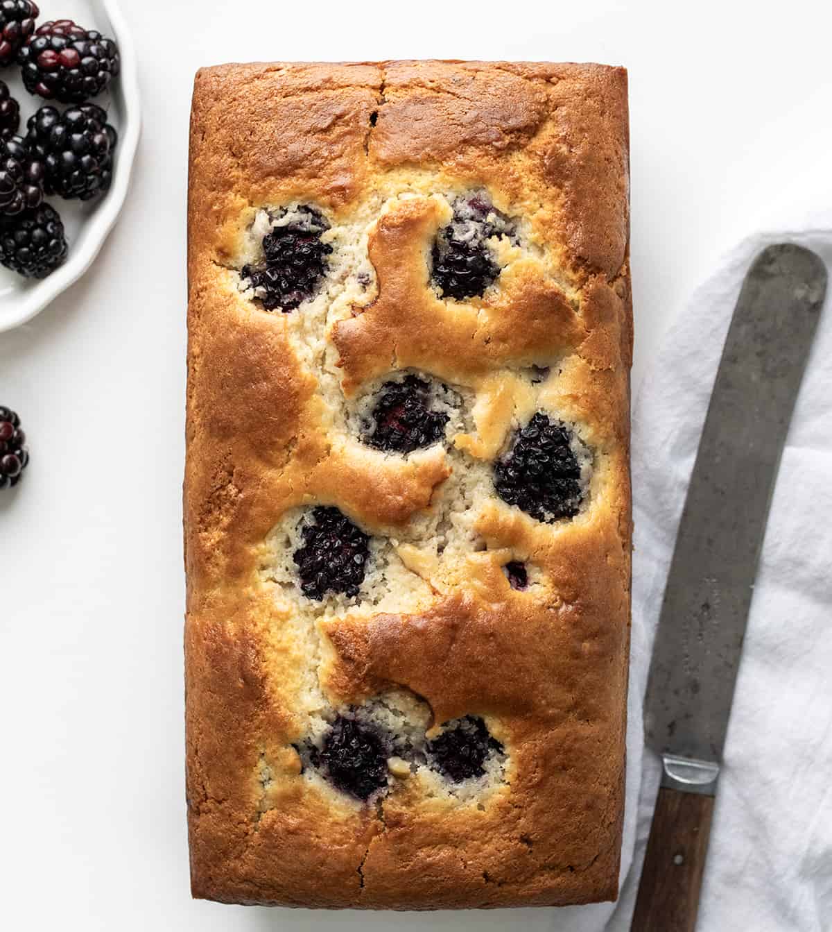 Blackberry Loaf is a soft and tender quick bread loaded with fresh blackberries and drizzled with an optional sweet glaze. Whether you're planning a casual brunch, afternoon tea, or simply a gathering with friends, this blackberry loaf fits the occasion perfectly! Plus, it is easy to make! I like to pair a slice with a cup of coffee for a delightful snack. Blackberry Loaf on a White Counter with Blackberries and Knife on a White Towel.