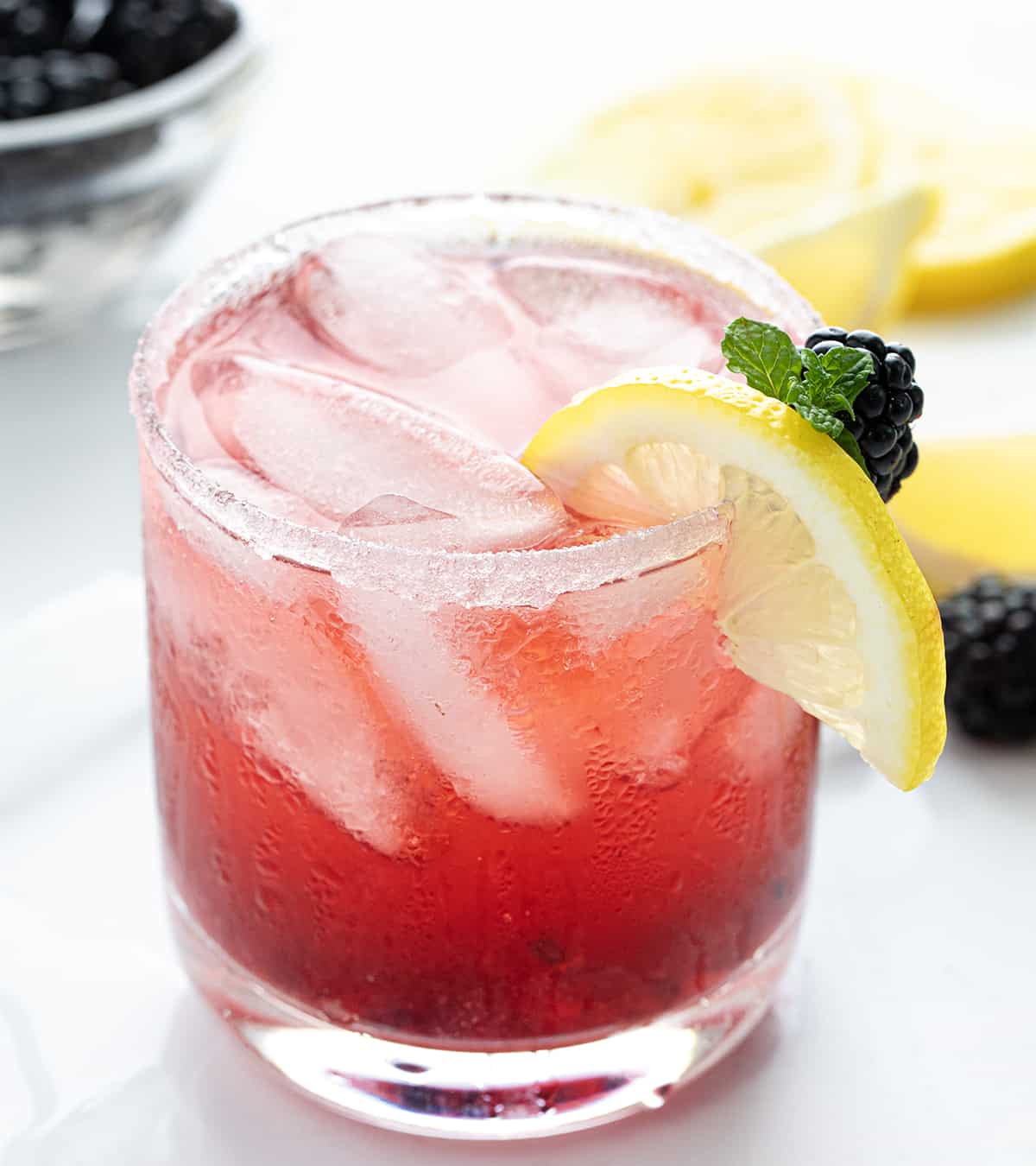 Blackberry Moscato Cocktail is a bubbly and refreshing alcoholic beverage made with fresh blackberries, lemon juice, sugar, lemon-lime soda, and Moscato d'Asti wine. This delightful drink is also known as Berry Breeze, Moscato Berry Smash, Lemonberry Fizz, and Blackberry Moscato Lemonade. No matter what you call it, you get a sweet, tangy, and fizzy concoction bursting with fruity flavors in every sip! One Blackberry Moscato Cocktail on a Counter with Lemons and Blackberries.