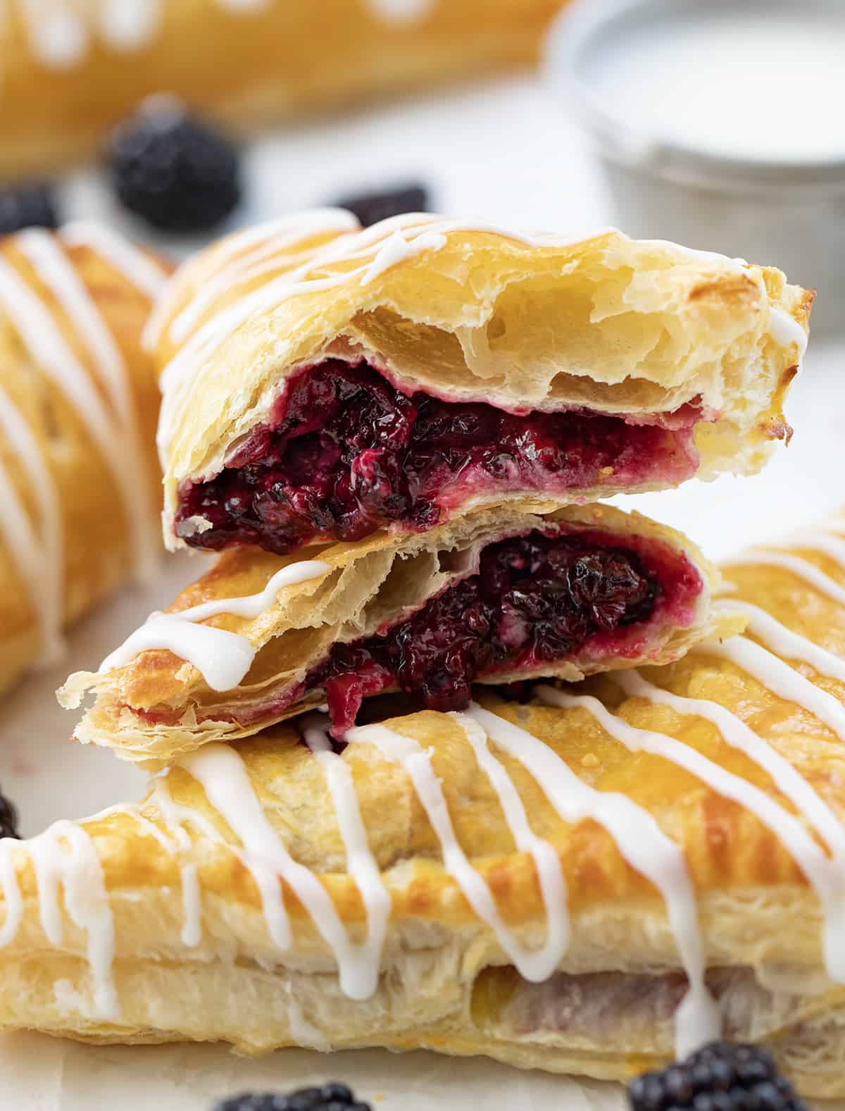 Blackberry Cream Cheese Turnovers are puff pastry squares filled with cream cheese and a homemade blackberry sauce, baked and drizzled with a sweet glaze. They are delicious as a breakfast pastry or a sweet treat for dessert. And, they can also be made in an air fryer! A Cut in Half Blackberry Cream Cheese Turnover on Top of a Whole Blackberry Cream Cheese Turnover on a White Counter.