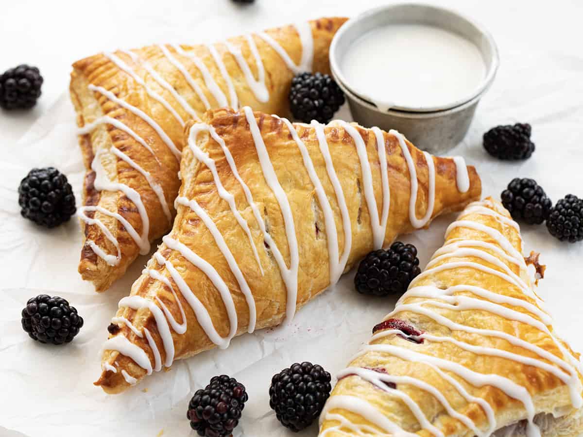Blackberry Cream Cheese Turnovers are puff pastry squares filled with cream cheese and a homemade blackberry sauce, baked and drizzled with a sweet glaze. They are delicious as a breakfast pastry or a sweet treat for dessert. And, they can also be made in an air fryer! Blackberry Cream Cheese Turnovers on a Pan with Glaze and a Few Blackberries on a White Counter.