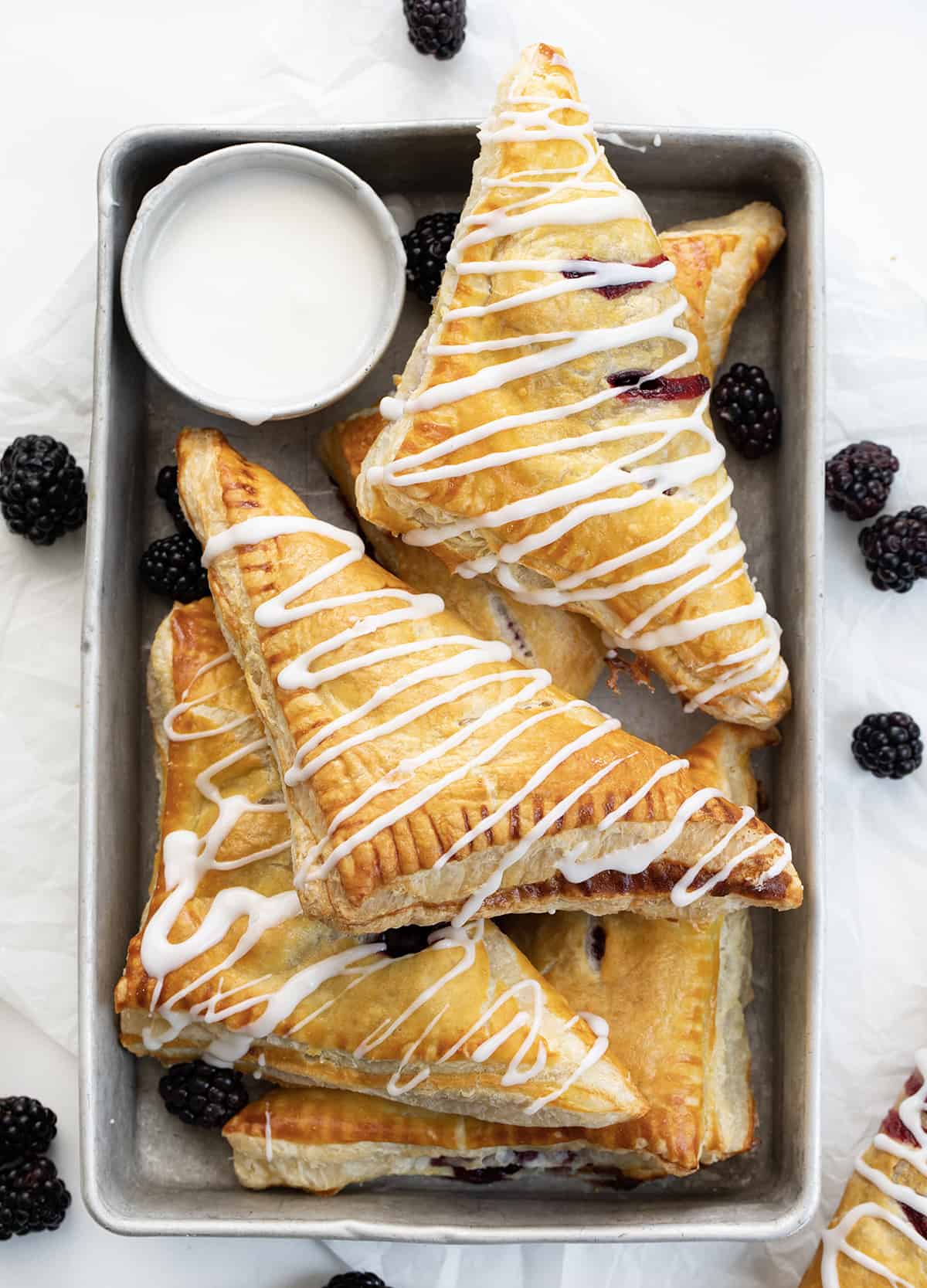 Blackberry Cream Cheese Turnovers are puff pastry squares filled with cream cheese and a homemade blackberry sauce, baked and drizzled with a sweet glaze. They are delicious as a breakfast pastry or a sweet treat for dessert. And, they can also be made in an air fryer! Blackberry Cream Cheese Turnovers on a Pan with Glaze and a Few Blackberries on a White Counter.