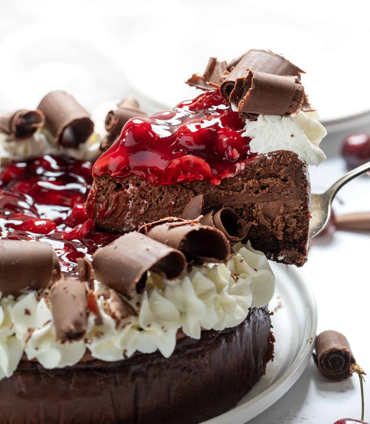 Black Forest Cheesecake is a rich chocolate cheesecake on top of an Oreo cookie crust, then topped with homemade cherry pie filling, chocolate curls, and whipped cream. It's a decadent dessert that brings together the delicious pairing of chocolate and cherries. Removing One Piece of Black Forest Cheesecake From the Serving Plate.