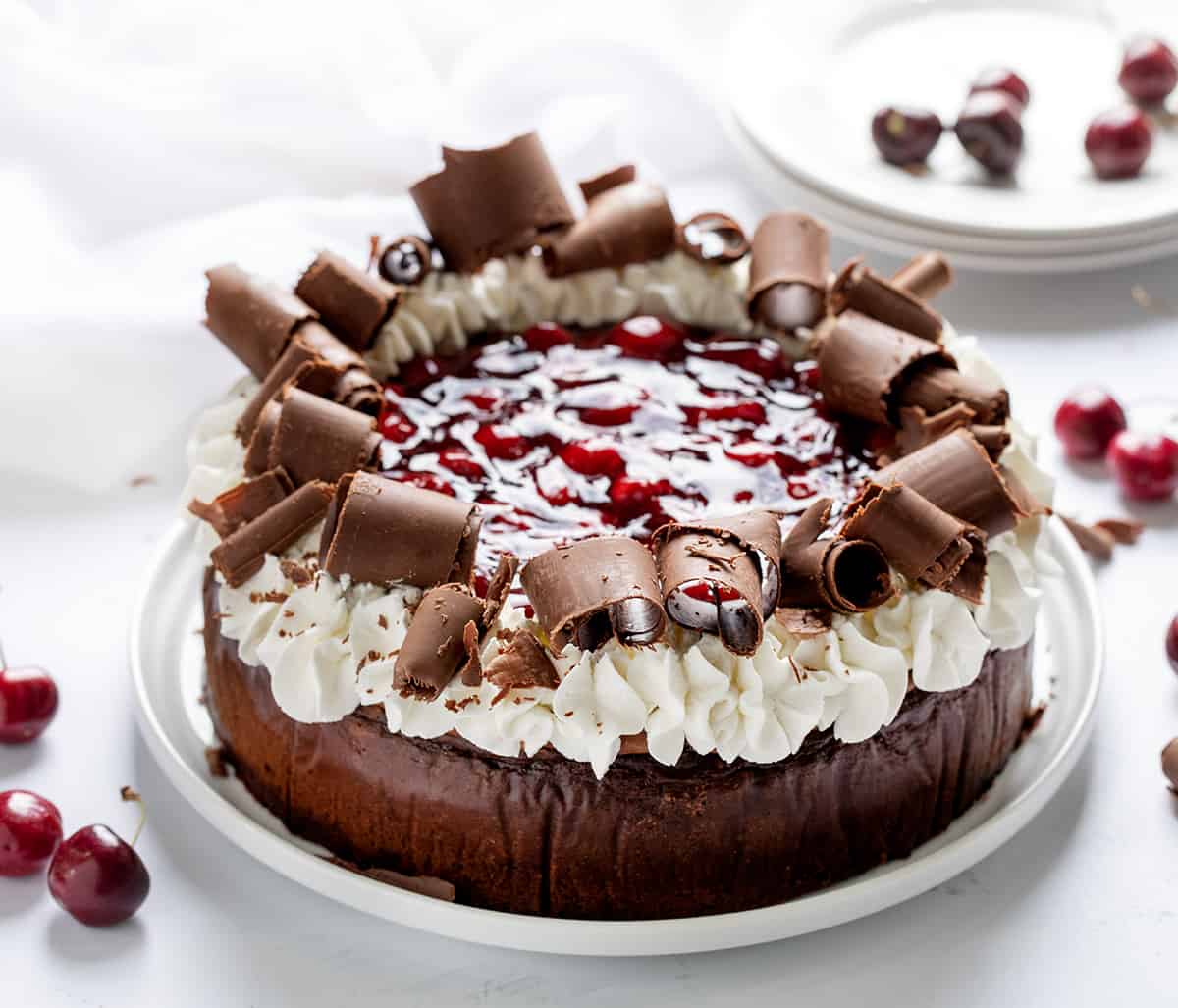 Black Forest Cheesecake is a rich chocolate cheesecake on top of an Oreo cookie crust, then topped with homemade cherry pie filling, chocolate curls, and whipped cream. It's a decadent dessert that brings together the delicious pairing of chocolate and cherries. Black Forest Cheesecake on a White Cake Plate on a White Surface and White Plates in the Background.