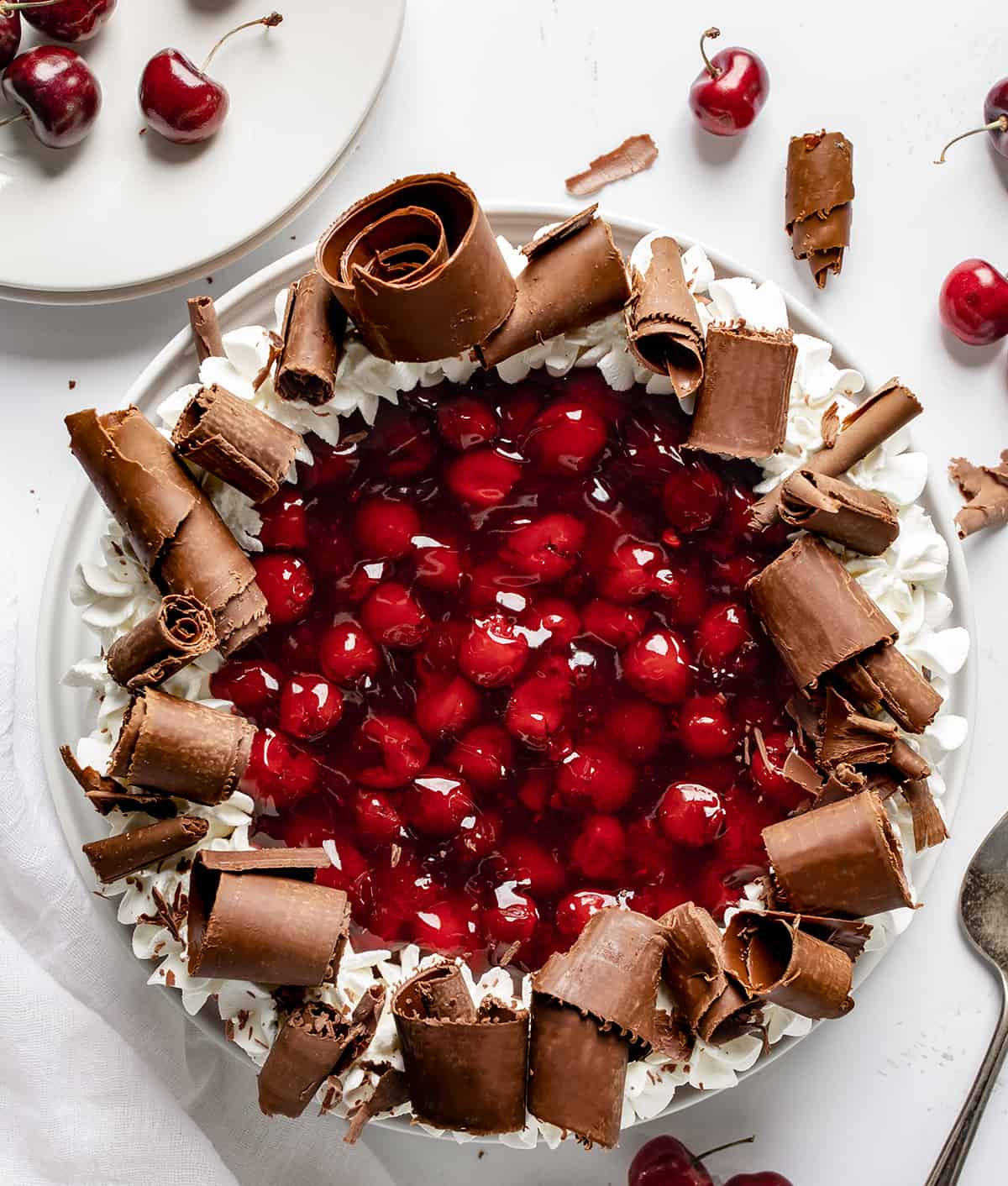 Black Forest Cheesecake is a rich chocolate cheesecake on top of an Oreo cookie crust, then topped with homemade cherry pie filling, chocolate curls, and whipped cream. It's a decadent dessert that brings together the delicious pairing of chocolate and cherries. Black Forest Cheesecake on a White Surface from Overhead with Some White Plates and Cherries Around.
