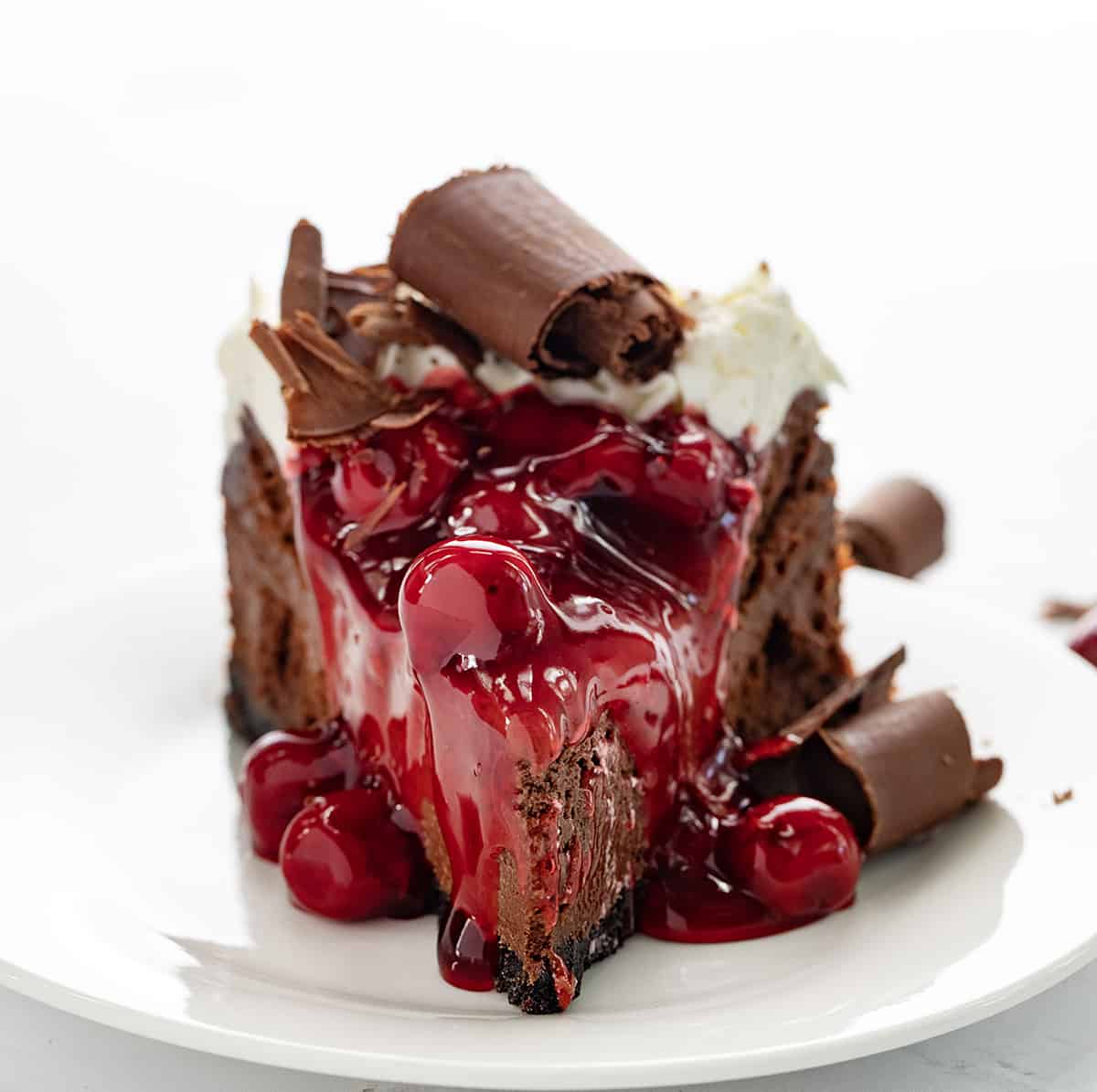 Black Forest Cheesecake is a rich chocolate cheesecake on top of an Oreo cookie crust, then topped with homemade cherry pie filling, chocolate curls, and whipped cream. It's a decadent dessert that brings together the delicious pairing of chocolate and cherries. One Piece of Black Forest Cheesecake on a White Plate and Backlit.