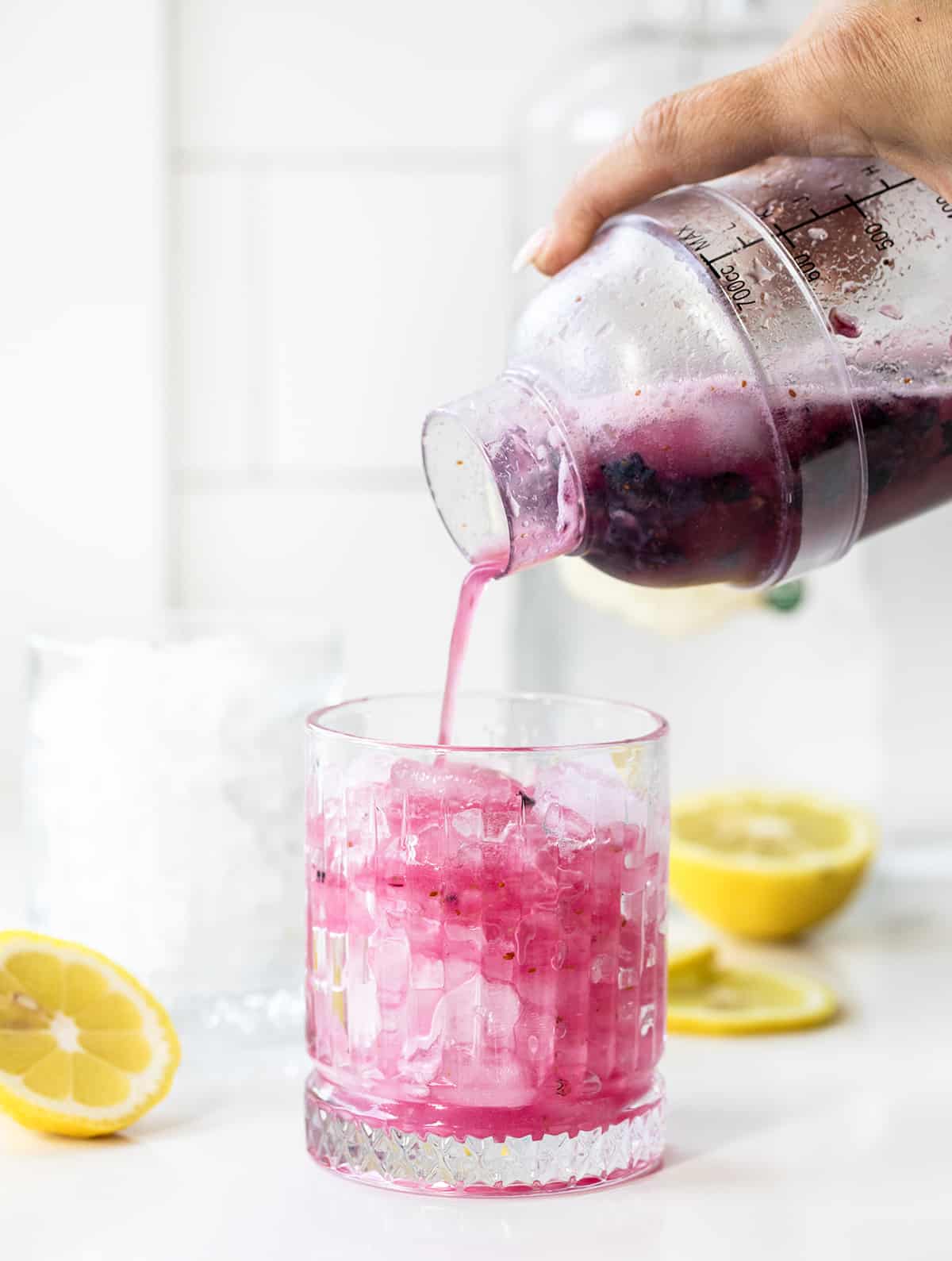 Spiked Blueberry Lemonade is a refreshing and vibrant cocktail made with fresh blueberries, simple syrup, freshly squeezed lemon juice, and vodka, topped off with club soda. Enjoy this fizzy alcoholic beverage over ice, garnished with lemon slices and blueberries for an elegant touch! Pouring Freshly Shaken Spiked Blueberry Lemonade from Shaker Into Glass with Ice in It on a White Counter with Lemons.