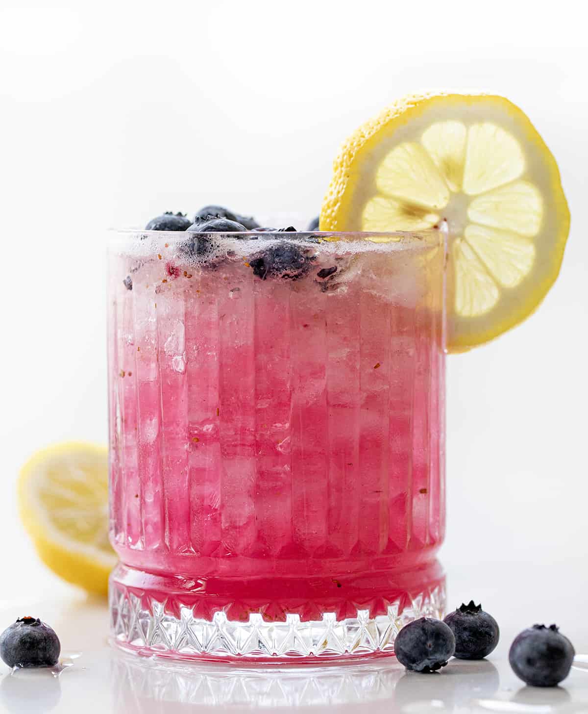Spiked Blueberry Lemonade is a refreshing and vibrant cocktail made with fresh blueberries, simple syrup, freshly squeezed lemon juice, and vodka, topped off with club soda. Enjoy this fizzy alcoholic beverage over ice, garnished with lemon slices and blueberries for an elegant touch! Glass of Spiked Blueberry Lemonade that is Backlit and Has a Lemon Wheel.