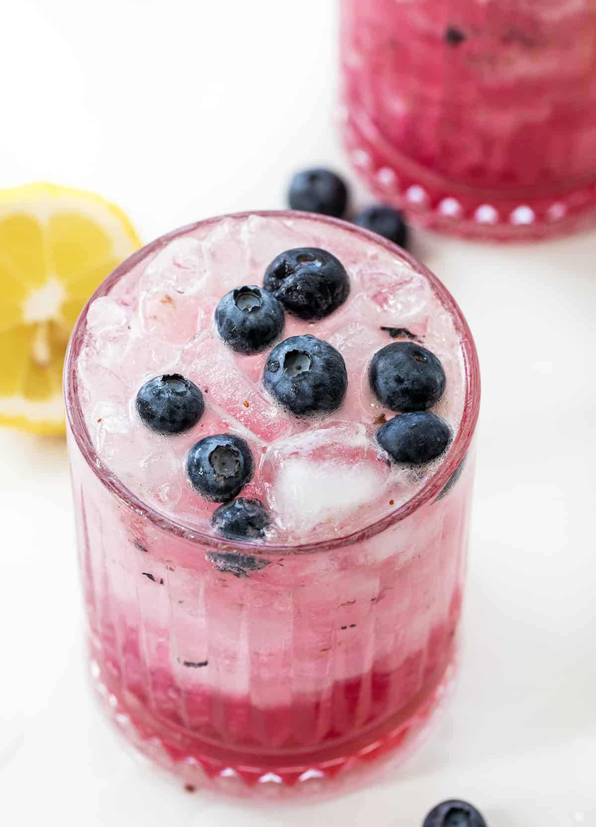 Spiked Blueberry Lemonade is a refreshing and vibrant cocktail made with fresh blueberries, simple syrup, freshly squeezed lemon juice, and vodka, topped off with club soda. Enjoy this fizzy alcoholic beverage over ice, garnished with lemon slices and blueberries for an elegant touch! Top of a Glass of Spiked Blueberry Lemonade Surrounded by Another Glass, Lemons, and Fresh Blueberries on a White Counter.