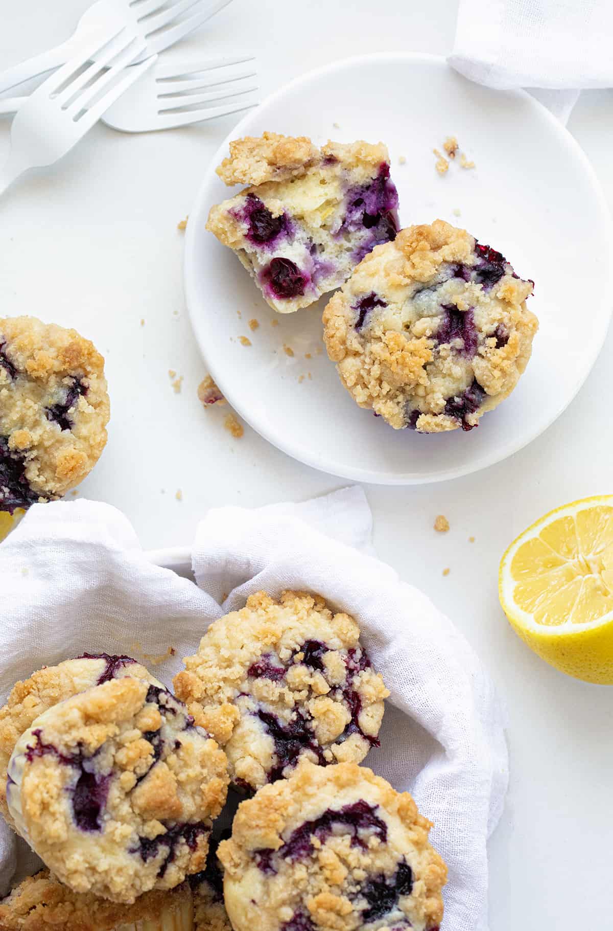Blueberry Lemon Crumb Muffins are moist and tender muffins loaded with fresh blueberries and lemon flavor, topped and baked with a crispy, buttery crumble. They are the perfect sweet snack, dessert, or breakfast! Blueberry Lemon Muffins in a Towel Lined Bowl Next to Muffins on a Plate with Fresh Lemons.