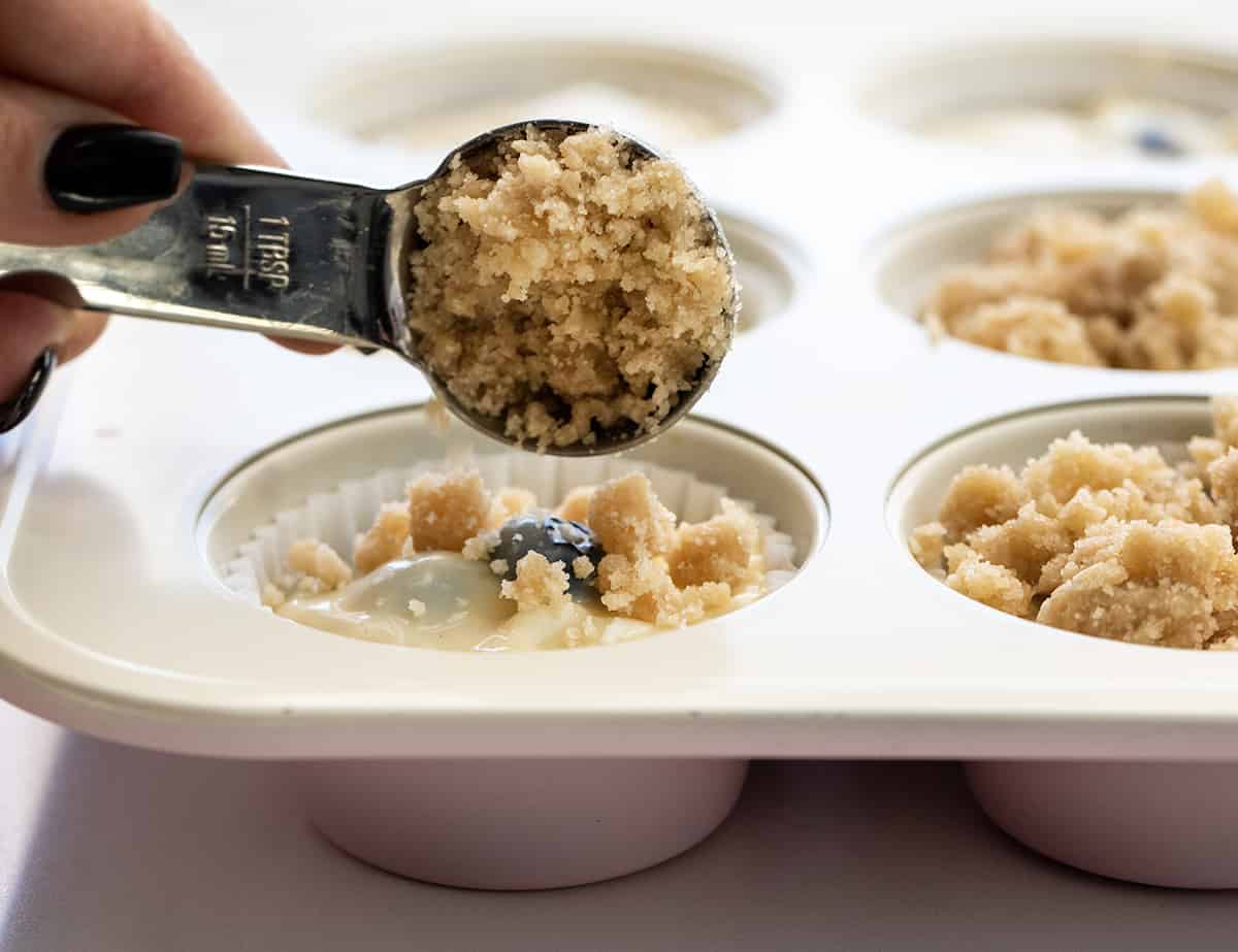 Blueberry Lemon Crumb Muffins are moist and tender muffins loaded with fresh blueberries and lemon flavor, topped and baked with a crispy, buttery crumble. They are the perfect sweet snack, dessert, or breakfast! Adding Crumb Topping to Blueberry Lemon Muffin Batter in a Pan.