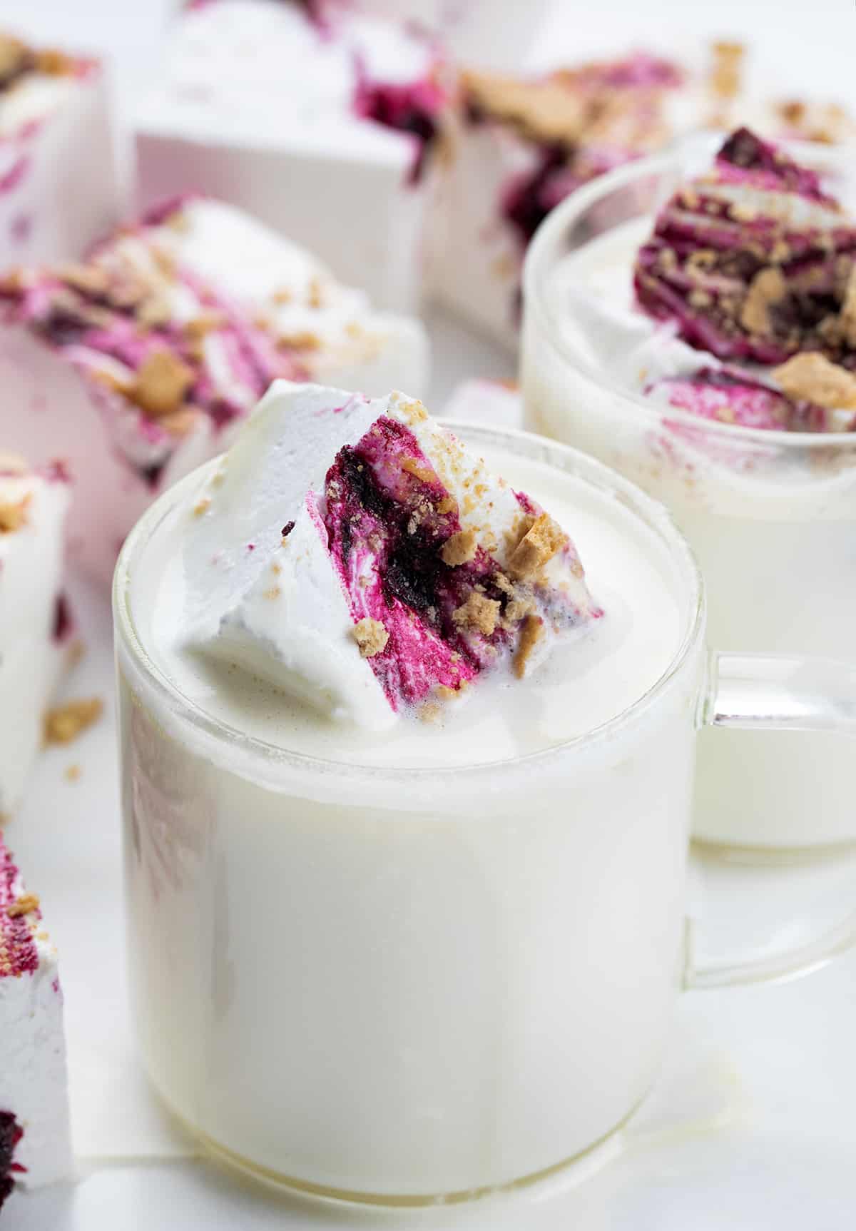 Blueberry Marshmallows are super fluffy homemade marshmallows marbled with blueberry syrup and topped with crushed graham crackers. I was going for a "Blueberry Cheesecake Marshmallow" and I think the addition of the graham crackers really works! The unique combination of flavors and textures elevates them (literally) to the level of gourmet marshmallows, making them a must-try for anyone who loves a sweet treat. Pieces of Gourmet Blueberry Marshmallows On a White Surface and Cut into Squares. Next to Cups of White Chocolate Hot Cocoa.