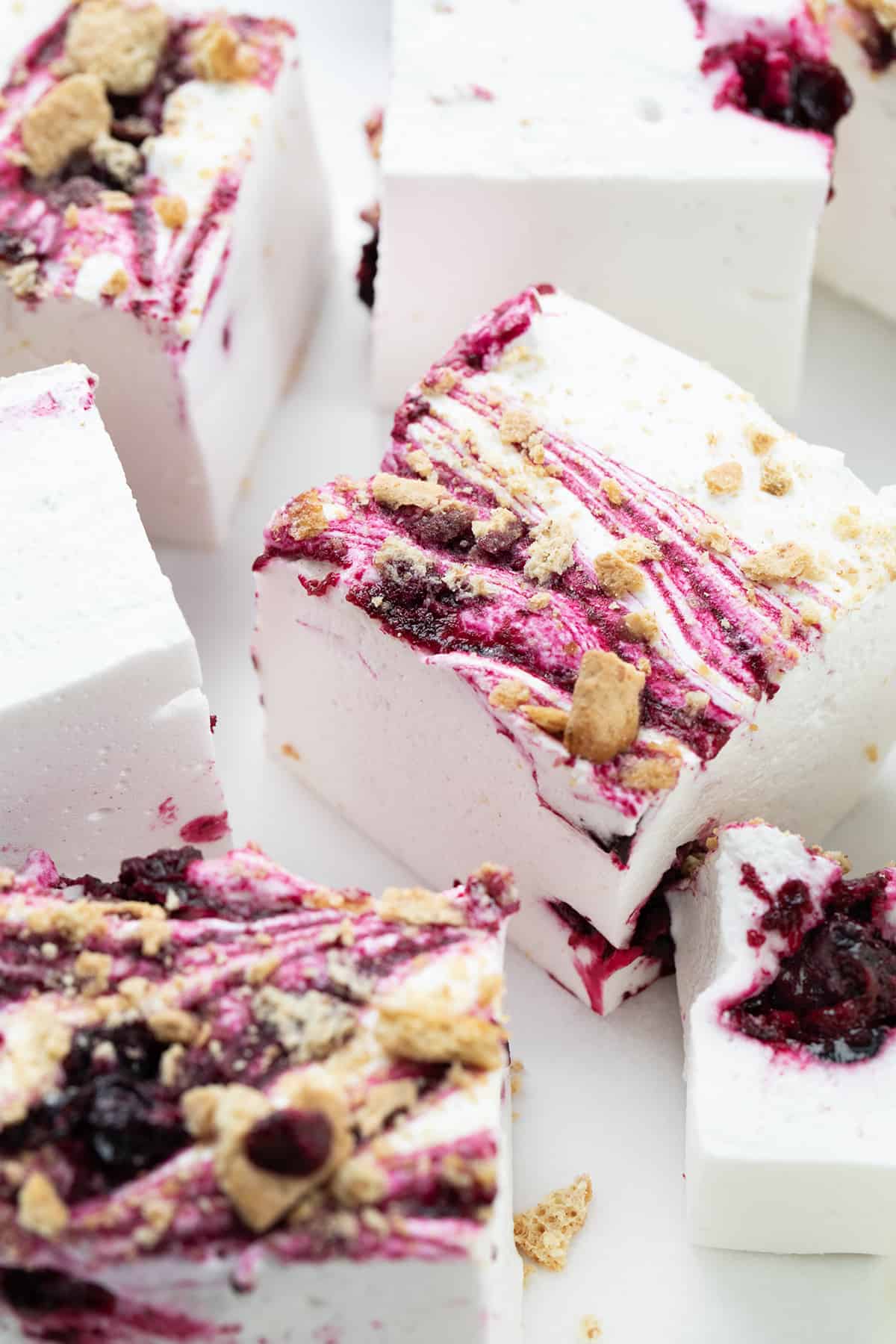 Blueberry Marshmallows are super fluffy homemade marshmallows marbled with blueberry syrup and topped with crushed graham crackers. I was going for a "Blueberry Cheesecake Marshmallow" and I think the addition of the graham crackers really works! The unique combination of flavors and textures elevates them (literally) to the level of gourmet marshmallows, making them a must-try for anyone who loves a sweet treat. Pieces of Gourmet Blueberry Marshmallows On a White Surface and Cut into Squares.