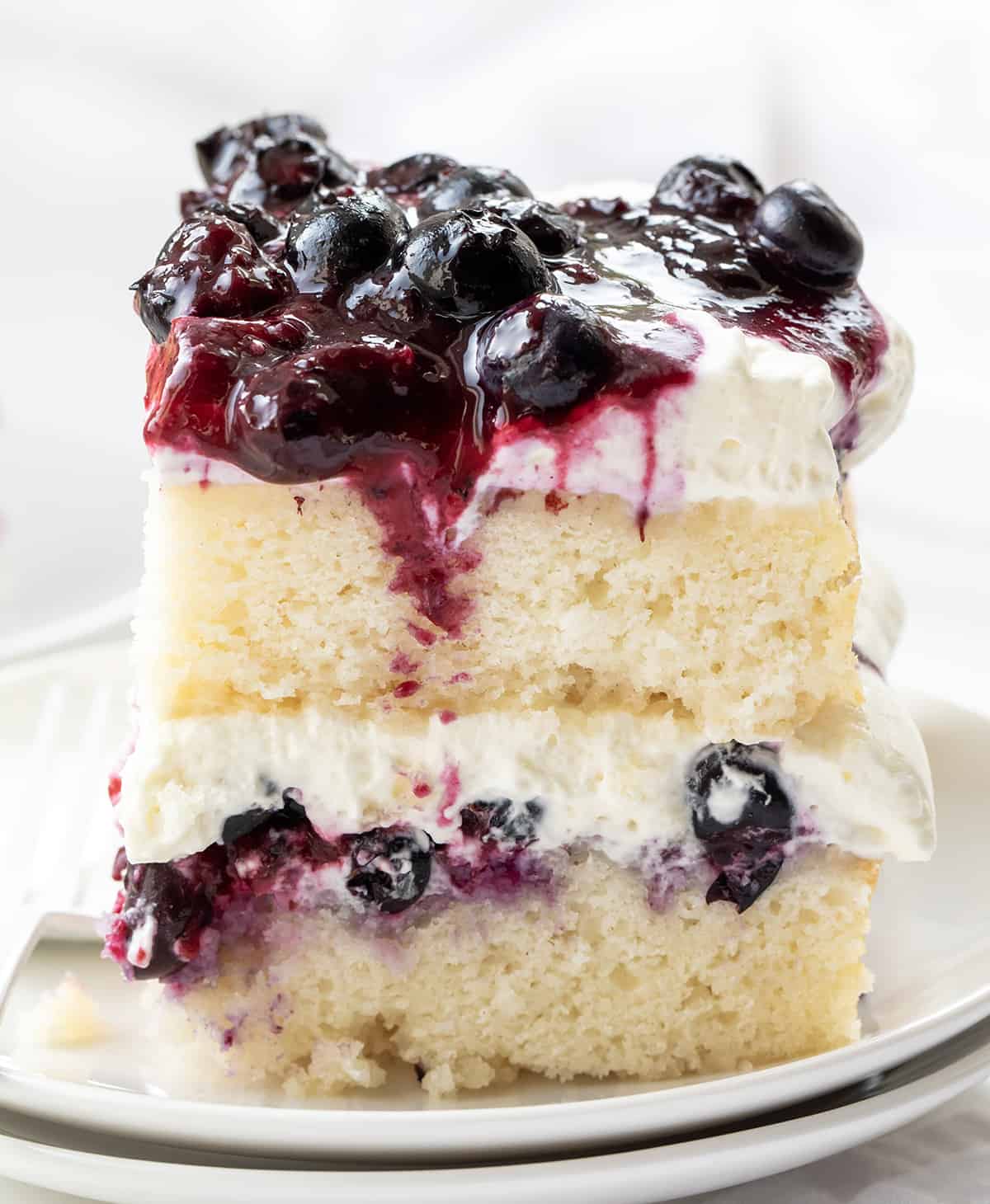 Sheet Pan Blueberry Shortcake Cake starts with a delicious white cake that is cut in half and layered with a homemade blueberry mixture and whipped cream cheese topping. It's a delightful dessert, combining the tender cake base with a sweet and tangy blueberry topping. The creamy whipped topping adds a luxurious and creamy element to the dessert, making it a perfect treat for any occasion. One Piece of Sheet Pan Blueberry Shortcake Cake on a White Plate.