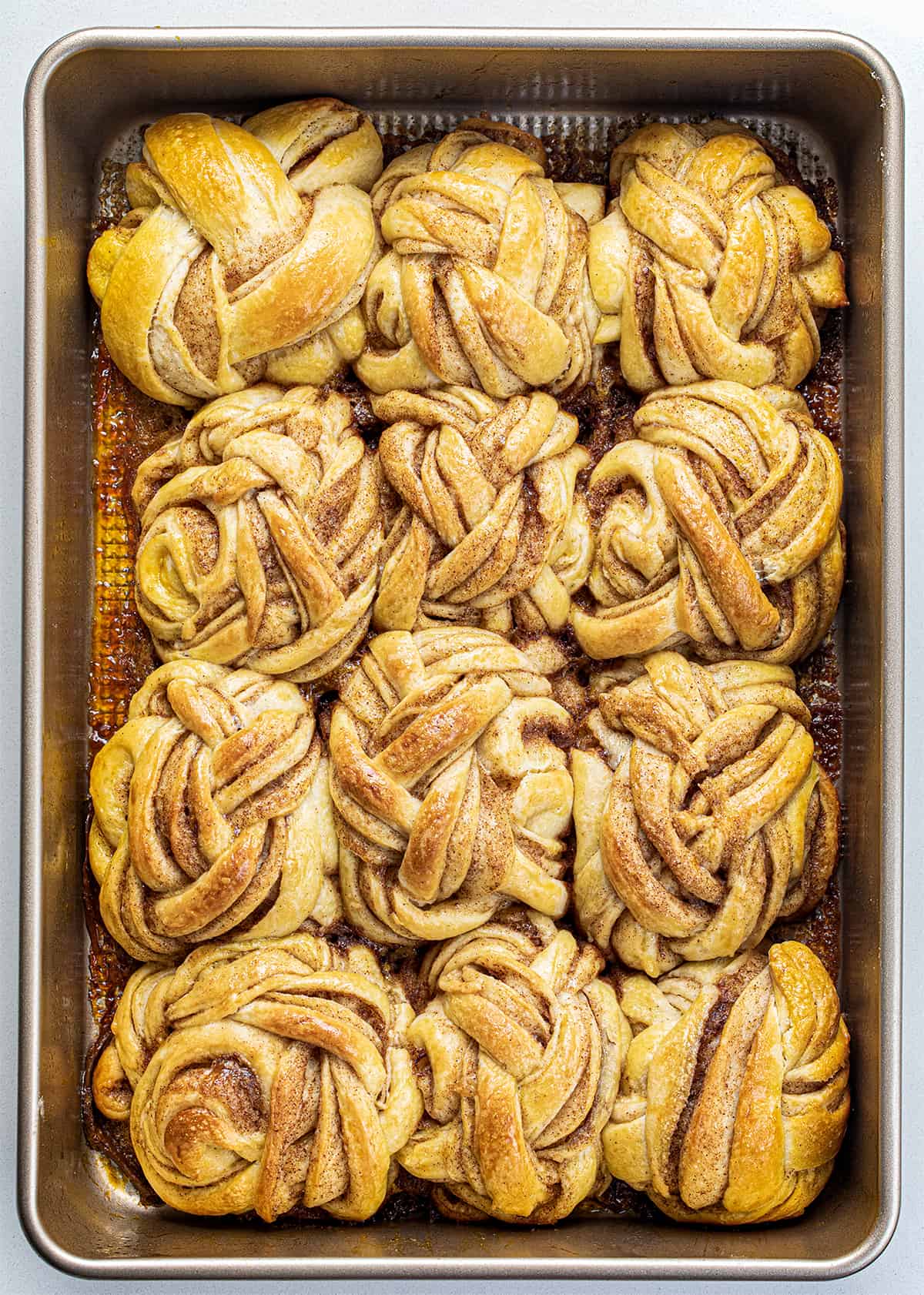 Braided Cinnamon Rolls are a delightful variation of the classic sweet rolls with a beautifully woven braided appearance, drizzled with a cream cheese glaze. They are so soft and the combination of cinnamon, brown sugar, and butter in each layer makes them so irresistible! They are wonderful without the glaze, too. You can also make these in an air fryer! Braided Cinnamon Rolls After Baking in a Pan from Overhead on a White Counter.