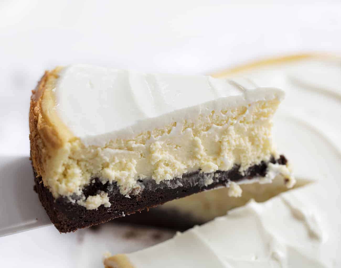 Brownie Bottom Cheesecake is a rich and creamy cheesecake that is layered on top of a sweet chocolate brownie mixture bottom, covered with a sweet sour cream topping! Cheesecake, Brownie Bottom Cheesecake, Brownie Cheesecake, Dessert, Baking, How to Make Brownie Bottom Cheesecake, How to Make Sour Cream Glaze, i am baker, iambaker Removing Piece of Brownie Bottom Cheesecake from Platter and Holding Up