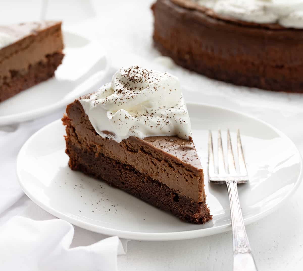 Brownie Bottom Chocolate Cheesecake is a homemade brownie crust topped with a creamy and decadent chocolate cheesecake. Then, homemade whipped cream is dolloped on to create a dessert that is sure to satisfy everyone's sweet tooth! It's like getting two desserts in one! Pieces of Brownie Bottom Chocolate Cheesecake on White Plates with Form.