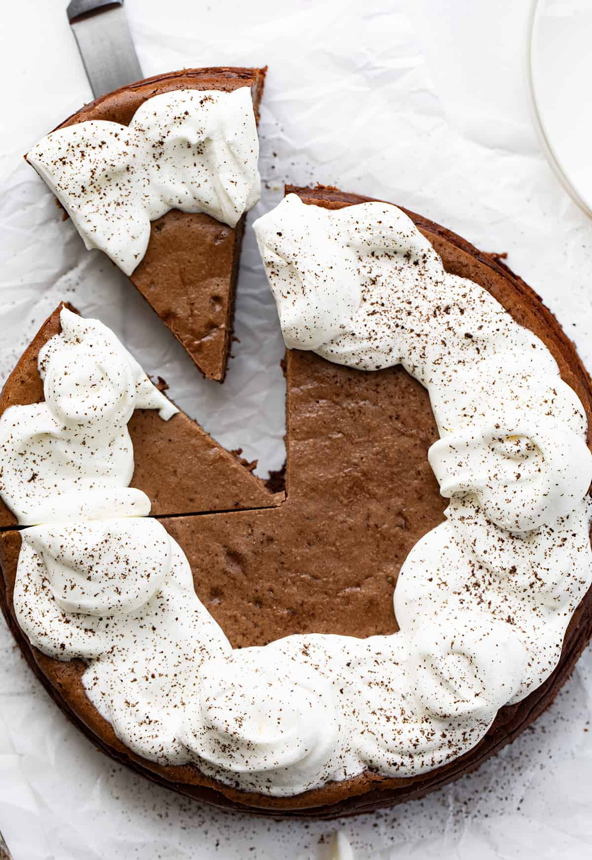 Brownie Bottom Chocolate Cheesecake is a homemade brownie crust topped with a creamy and decadent chocolate cheesecake. Then, homemade whipped cream is dolloped on to create a dessert that is sure to satisfy everyone's sweet tooth! It's like getting two desserts in one! Whole Brownie Bottom Chocolate Cheesecake on White Parchment with Homemade Whipped Topping and Dusted with Chocolate with One Piece Being Removed.