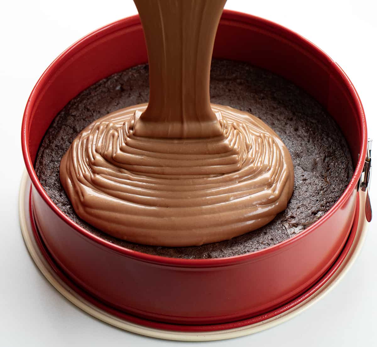 Brownie Bottom Chocolate Cheesecake is a homemade brownie crust topped with a creamy and decadent chocolate cheesecake. Then, homemade whipped cream is dolloped on to create a dessert that is sure to satisfy everyone's sweet tooth! It's like getting two desserts in one! Pouring Chocolate Cheesecake Over Baked Brownie to Make Brownie Bottom Chocolate Cheesecake.