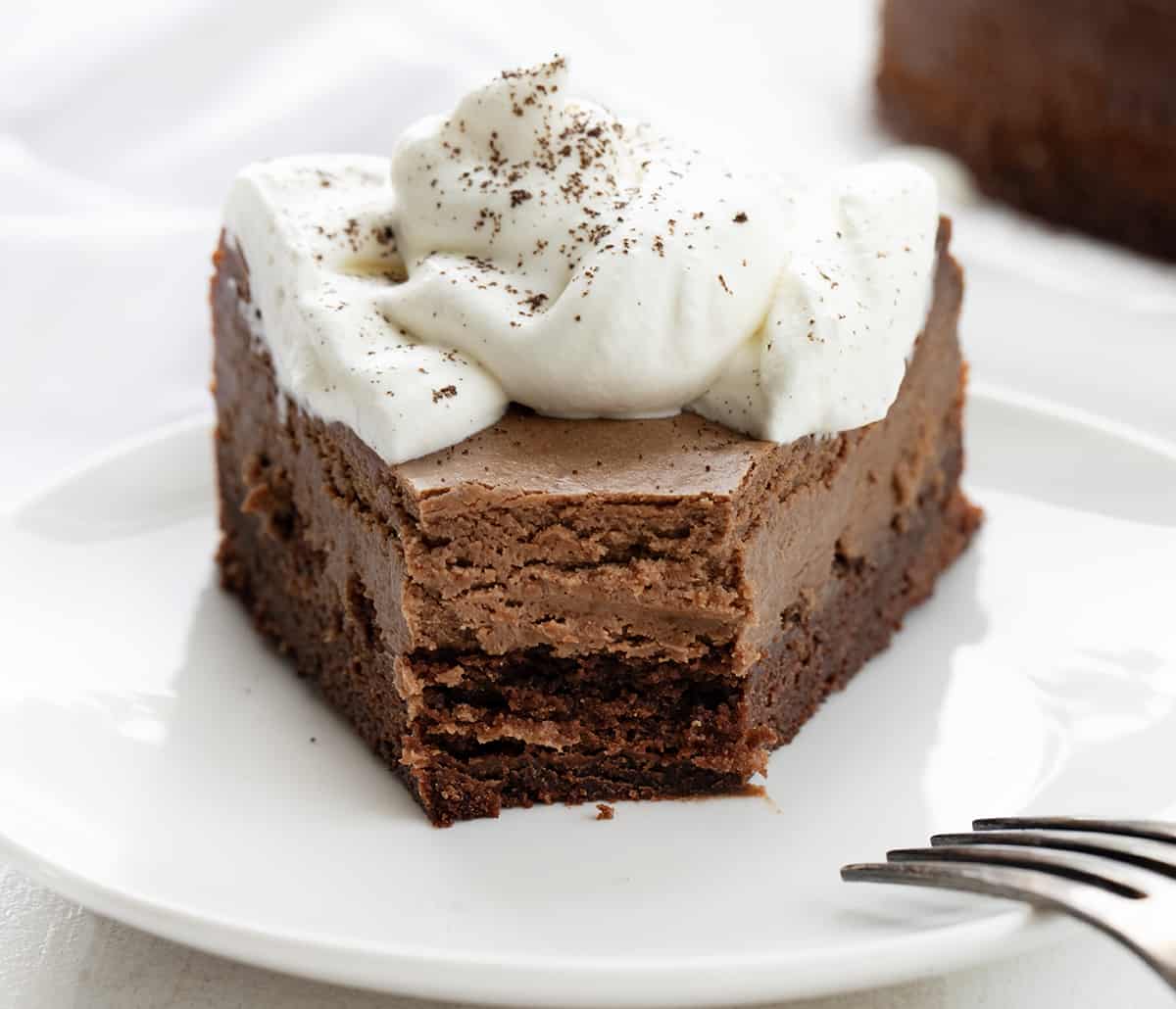 Brownie Bottom Chocolate Cheesecake is a homemade brownie crust topped with a creamy and decadent chocolate cheesecake. Then, homemade whipped cream is dolloped on to create a dessert that is sure to satisfy everyone's sweet tooth! It's like getting two desserts in one! Piece of Brownie Bottom Chocolate Cheesecake on a White Plate with Bite Removed Showing Inside Texture.