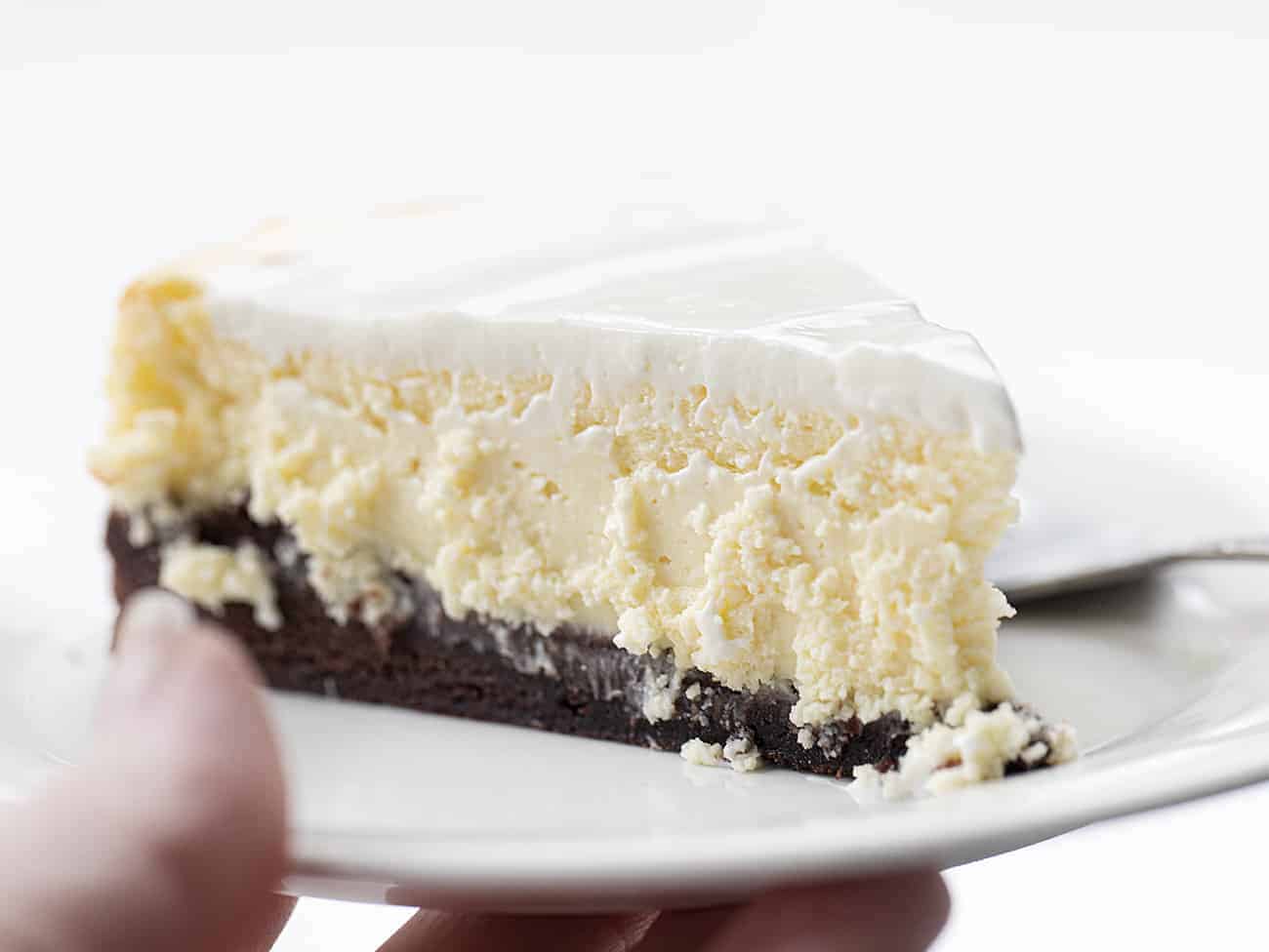 Brownie Bottom Cheesecake is a rich and creamy cheesecake that is layered on top of a sweet chocolate brownie mixture bottom, covered with a sweet sour cream topping! Cheesecake, Brownie Bottom Cheesecake, Brownie Cheesecake, Dessert, Baking, How to Make Brownie Bottom Cheesecake, How to Make Sour Cream Glaze, i am baker, iambaker Hand Holding White Plate with Brownie Bottom Cheesecake Recipe with Sour Cream Frosting Topping and Fork Resting Next to It