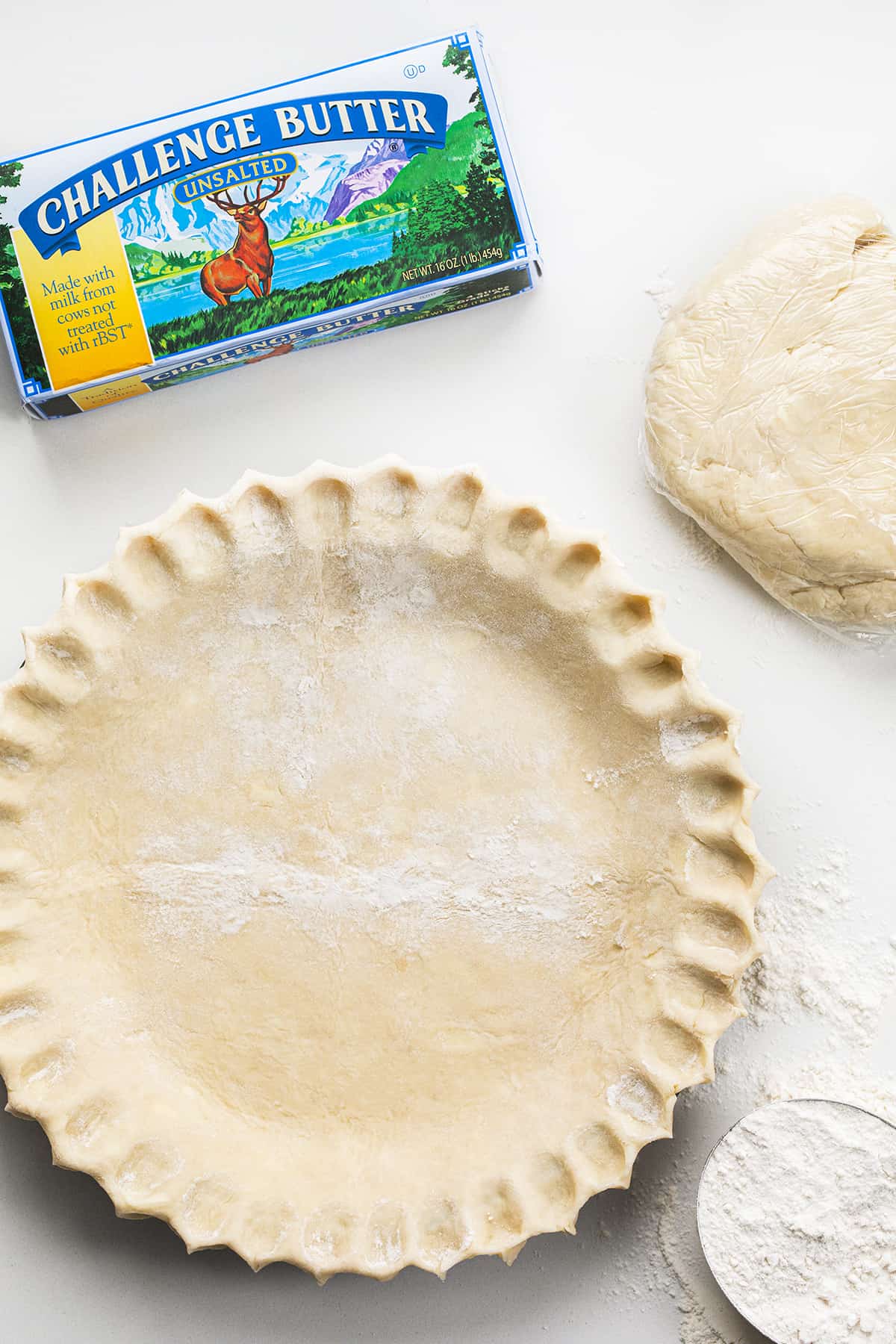 Do you want the best no-fail ALL BUTTER Pie crust? Professional bakers agree, this one tastes the best! Pie Crust in a Pie that has Been Crimped Before Baking Next to Flour, Butter, and an Additional Pie Crust in Plastic Wrap.