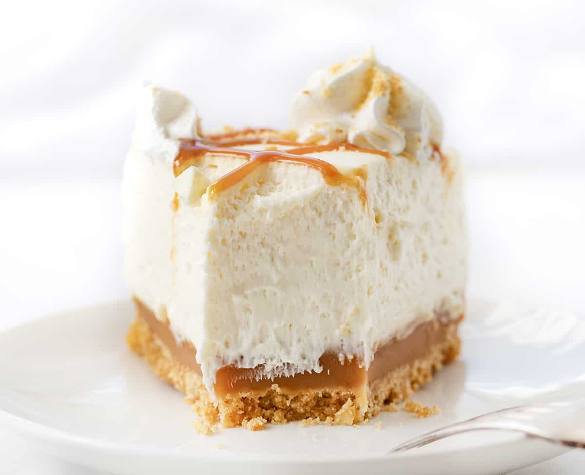 Caramel Cheesecake is a no-bake dessert with a buttery shortbread crust layered with a glorious layer of homemade caramel and a smooth, creamy cheesecake filling. Then, it's topped with more crumbled shortbread cookies, dollops of whipped cream, and caramel for a sweet, rich, and tangy treat! Piece of Caramel Cheesecake with Bite Removed on a White Plate.