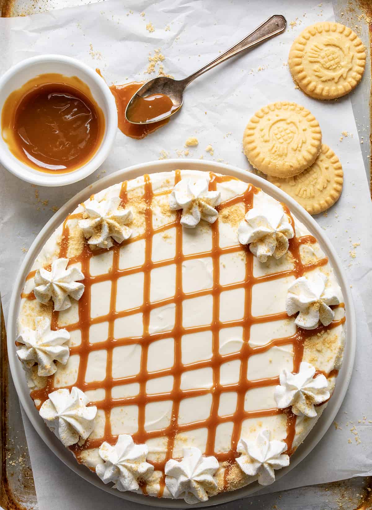 Caramel Cheesecake is a no-bake dessert with a buttery shortbread crust layered with a glorious layer of homemade caramel and a smooth, creamy cheesecake filling. Then, it's topped with more crumbled shortbread cookies, dollops of whipped cream, and caramel for a sweet, rich, and tangy treat! Caramel Cheesecake from Overhead with some Shortbread Cookies and Caramel Nearby.