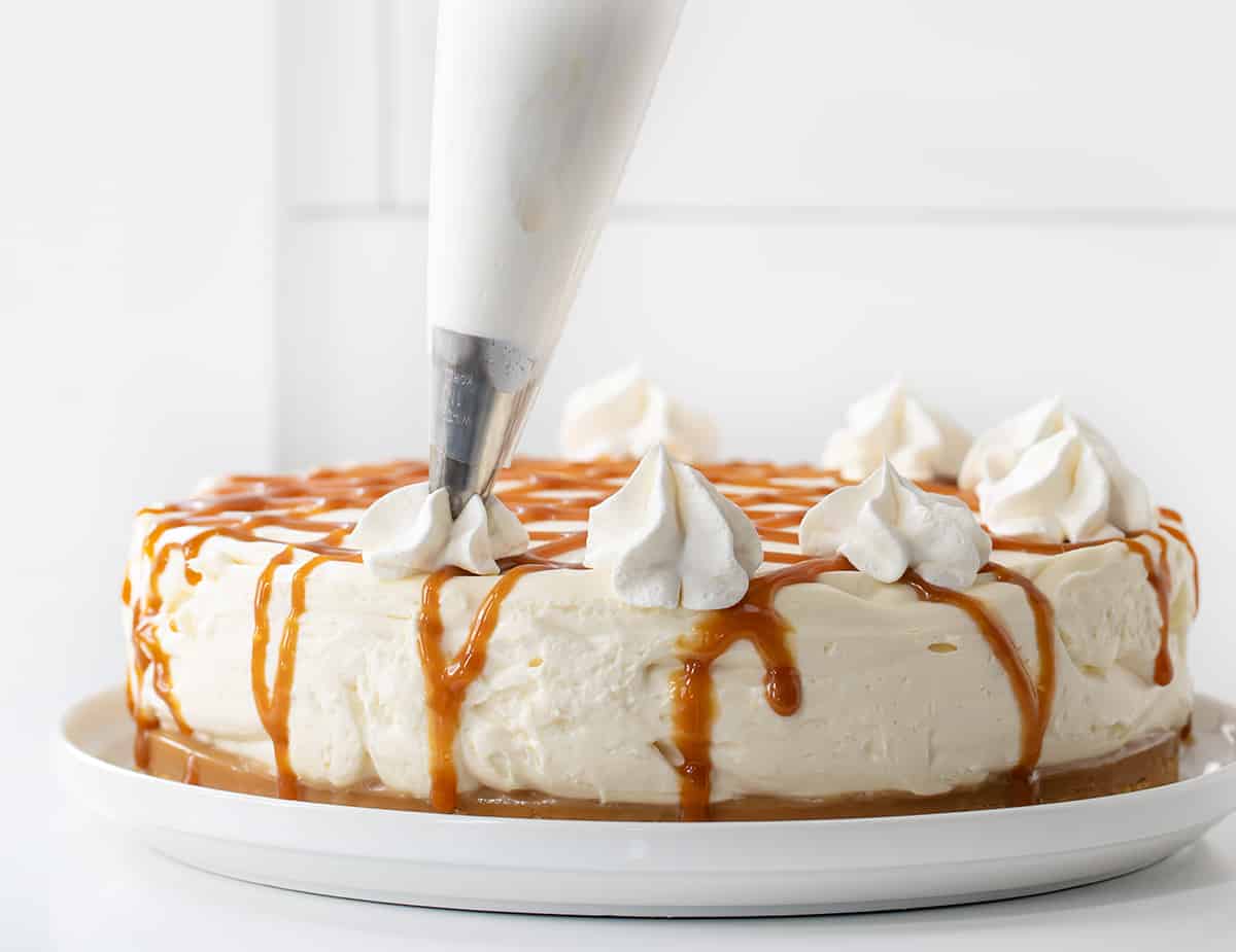 Caramel Cheesecake is a no-bake dessert with a buttery shortbread crust layered with a glorious layer of homemade caramel and a smooth, creamy cheesecake filling. Then, it's topped with more crumbled shortbread cookies, dollops of whipped cream, and caramel for a sweet, rich, and tangy treat! Adding Dollops of Whipped Topping to a Caramel Cheesecake on a White Plate.