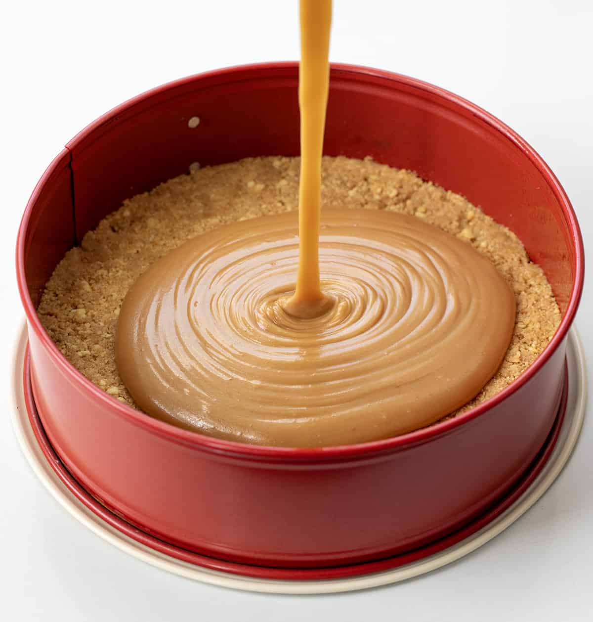 Caramel Cheesecake is a no-bake dessert with a buttery shortbread crust layered with a glorious layer of homemade caramel and a smooth, creamy cheesecake filling. Then, it's topped with more crumbled shortbread cookies, dollops of whipped cream, and caramel for a sweet, rich, and tangy treat! Pouring Caramel into Springform Pan.