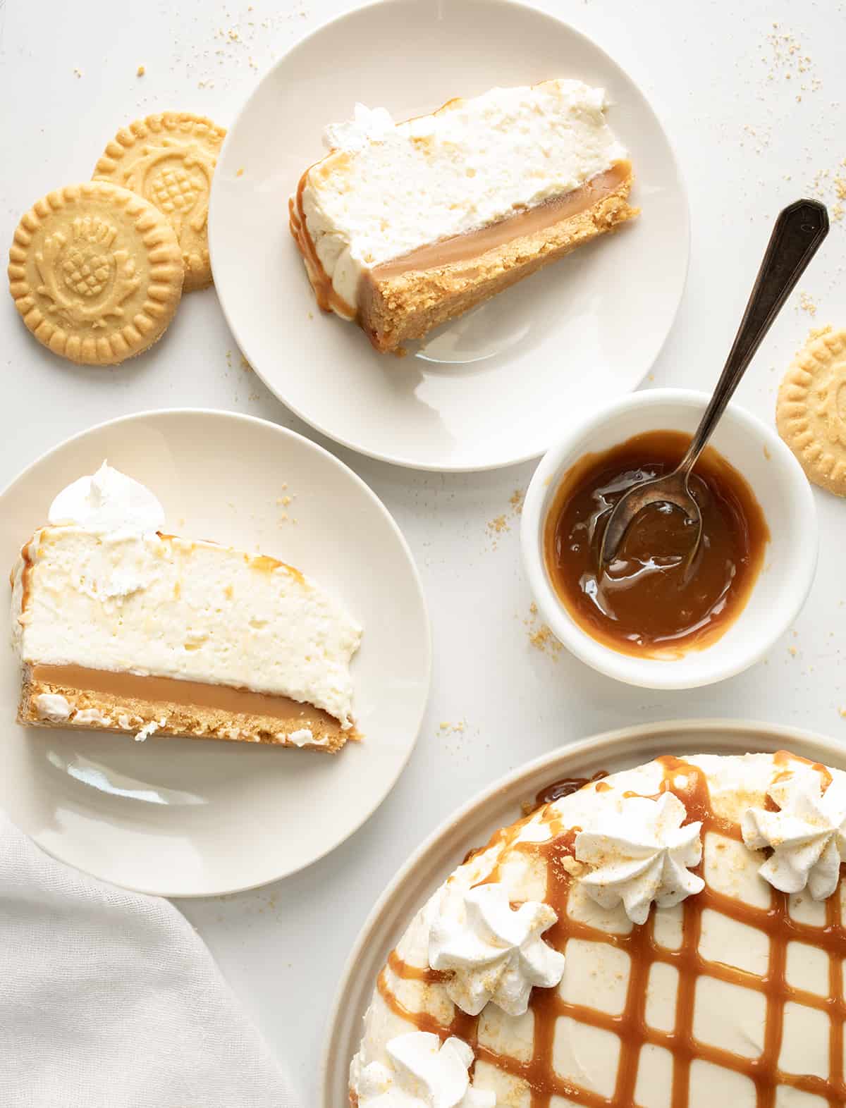 Caramel Cheesecake is a no-bake dessert with a buttery shortbread crust layered with a glorious layer of homemade caramel and a smooth, creamy cheesecake filling. Then, it's topped with more crumbled shortbread cookies, dollops of whipped cream, and caramel for a sweet, rich, and tangy treat! Pieces of Caramel Cheesecake on White Plates with a Bowl a Caramel Next to a Caramel Cheesecake.