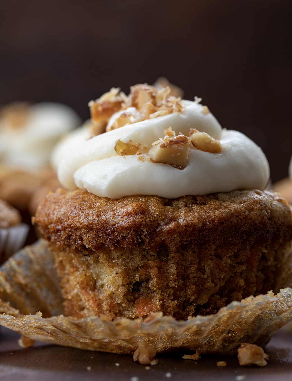 Carrot Cake Cupcakes are tender cupcakes loaded with carrots, walnuts, and pineapple that are topped with the best cream cheese frosting! The cupcakes give you all of the flavors of a cream cheese-frosted carrot cake but in a cupcake! The top is slightly crispy and the inside is a soft, tender crumb that is loaded with flavor. Be sure to have them on hand whenever you are craving carrot cake! Close up of a Carrot Cake Cupcake with Wrapper Peeled Back and Candied Walnuts on Top of the Cream Cheese Frosting.