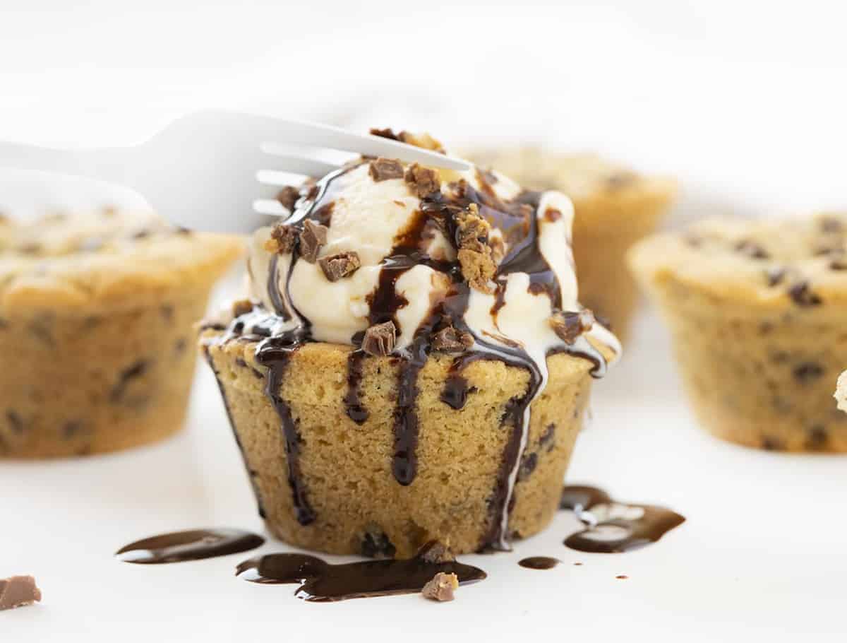 Chocolate Chip Cookie Cups is chocolate chip cookie dough baked in a muffin tin with a surprise mini Reese's Peanut Butter Cup nestled inside! Chocolate Chip Cookie Cups, Mini Chocolate Chip Cookie Cups, Cookie Cups, Cookies in Muffin Pan, Mini Chocolate Chip Cookie Recipe, Chocolate Chip Cookies Stuffed with Reese's, Reese's Chocolate Chip Cookies, Baking, Cookies, Cookie Recipes, CookieCups, i am baker, iambaker Fork Cutting into a Chocolate Chip Cookie Cup Stuffed with Reese's Peanut Butter Cup and Drizzled with Hot Fudge