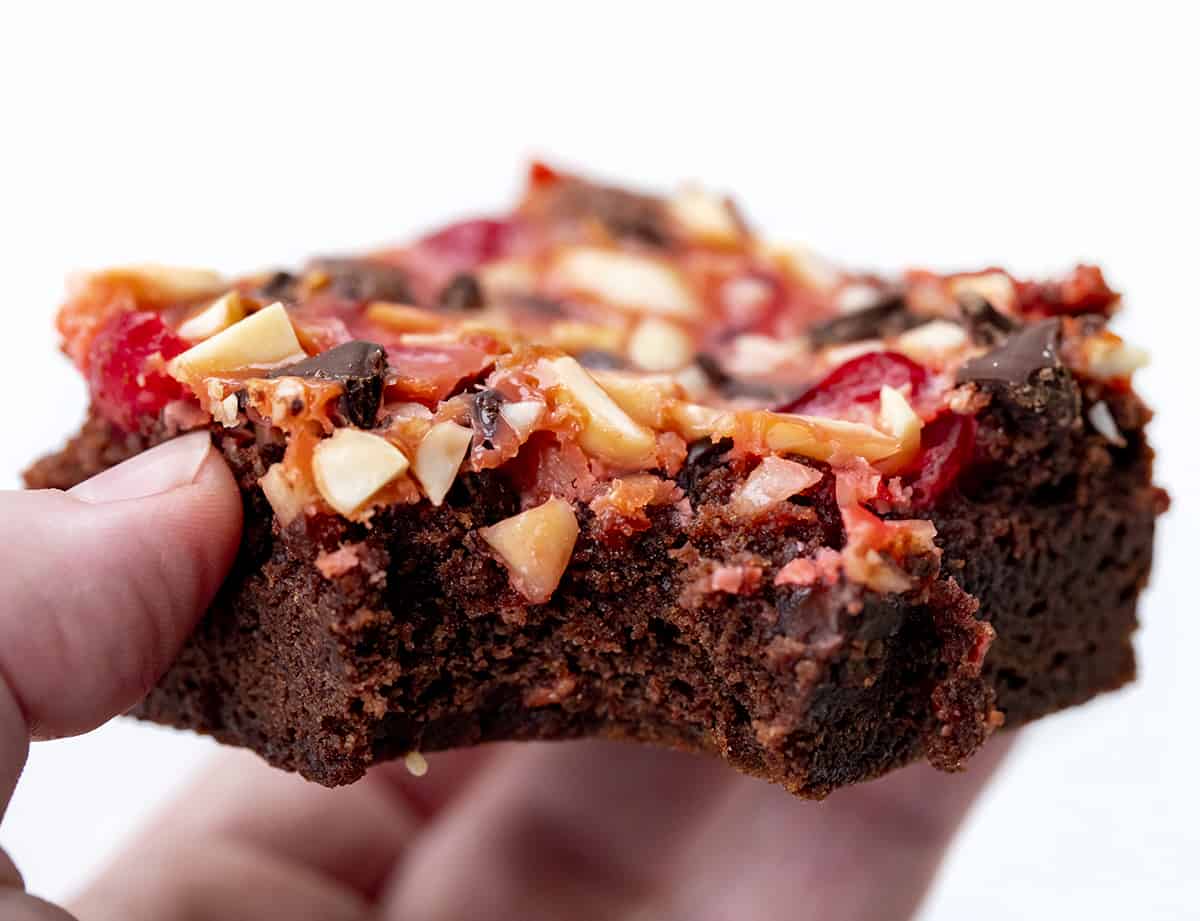 Cherry Chocolate Magic Brownies start with a brownie base topped and baked with maraschino cherries, chocolate chips, almonds, and sweetened condensed milk. It's like enjoying chocolate-covered cherries in a brownie! Hand holding bit into Cherry Chocolate Magic Brownies.