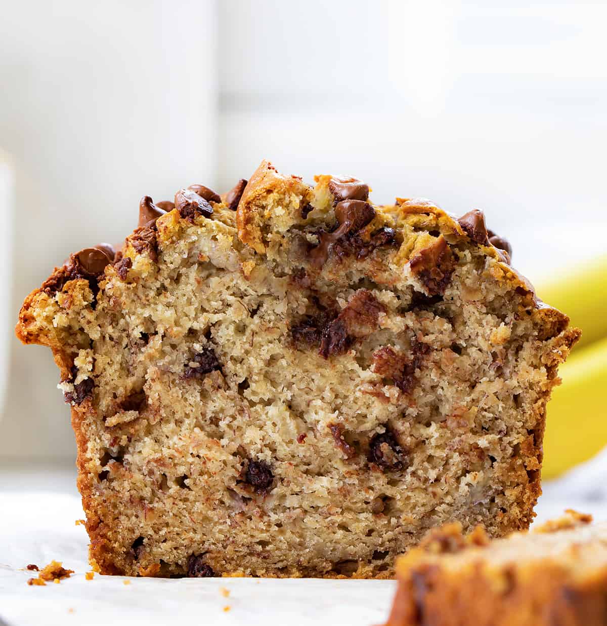 Chocolate Chip Banana Bread is a moist, delicious treat that is perfect for breakfast, brunch, or a snack. It is made with bananas and chocolate chips and is sure to please everyone. I make mine mile-high and just love how it rises in the pan! Cut into Loaf of Chocolate Chip Banana Bread on a White Counter with Bananas in the Background.