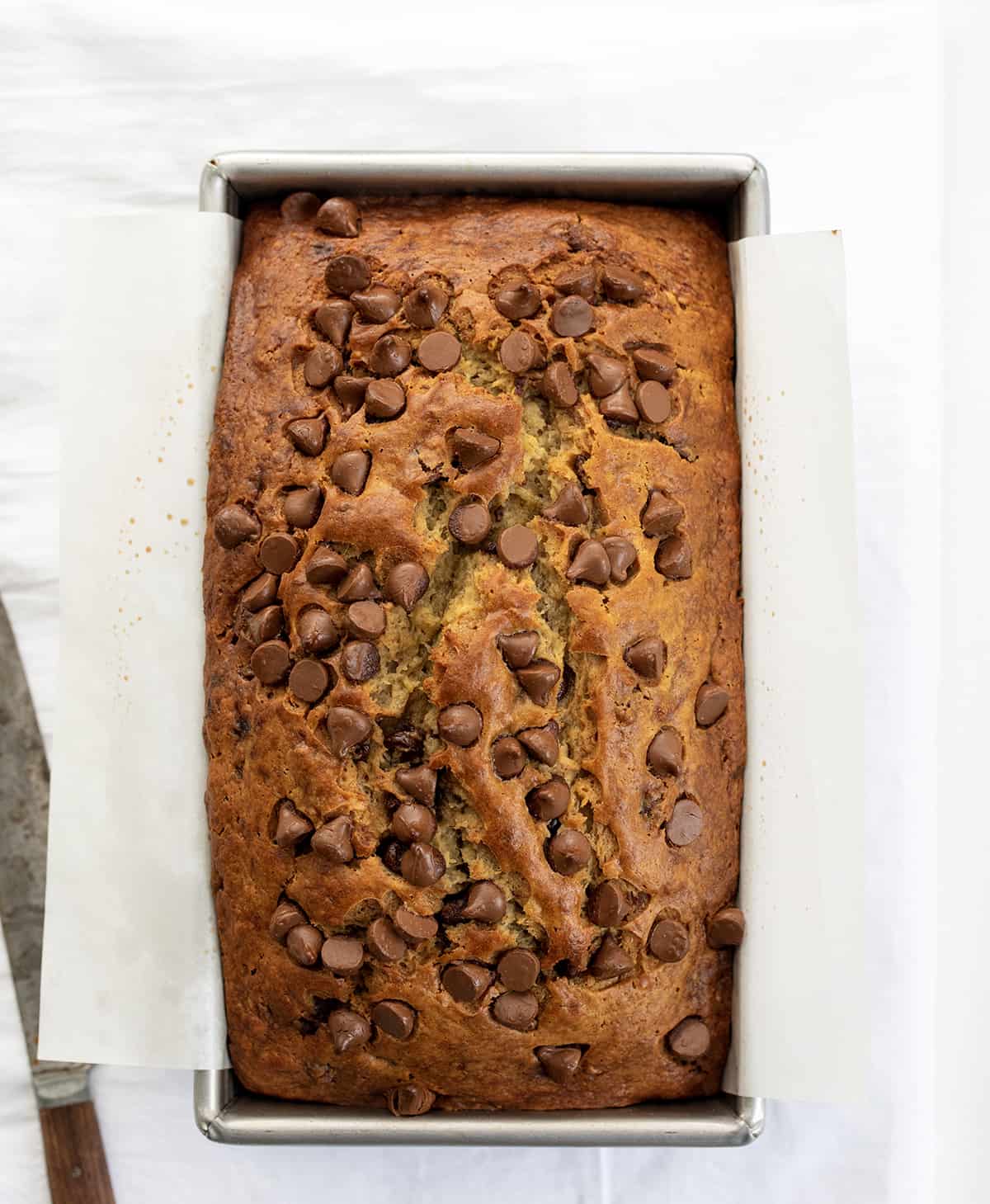 Chocolate Chip Banana Bread is a moist, delicious treat that is perfect for breakfast, brunch, or a snack. It is made with bananas and chocolate chips and is sure to please everyone. I make mine mile-high and just love how it rises in the pan! Loaf of Chocolate Chip Banana Bread on a White Counter with a Bread Knife.