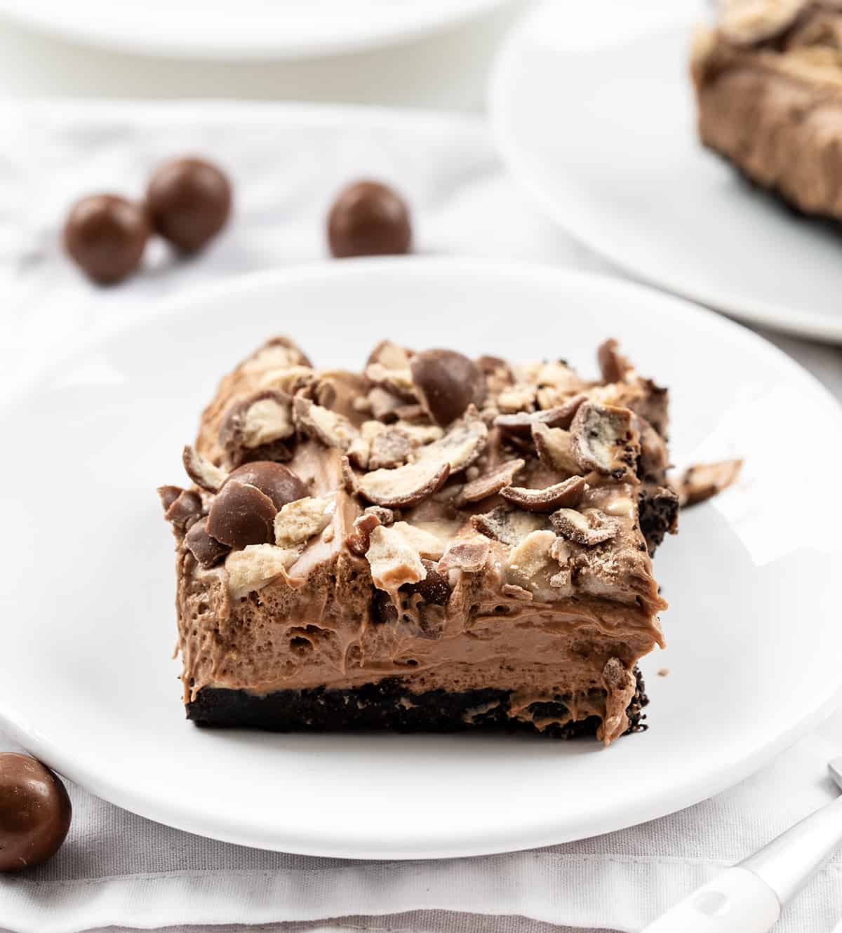 Chocolate Malt Shoppe Bars are creamy chocolatey bars with added chocolate malt powder over an Oreo cookie crust and topped with crushed malted milk ball (whopper) candies. This no-bake treat is like enjoying your favorite chocolate malted milkshake but in a dessert bar without any ice cream. Let each bite take you back to the nostalgic charm of your favorite malt shop! Plates of Chocolate Malt Shoppe Bars with Whoppers Around.