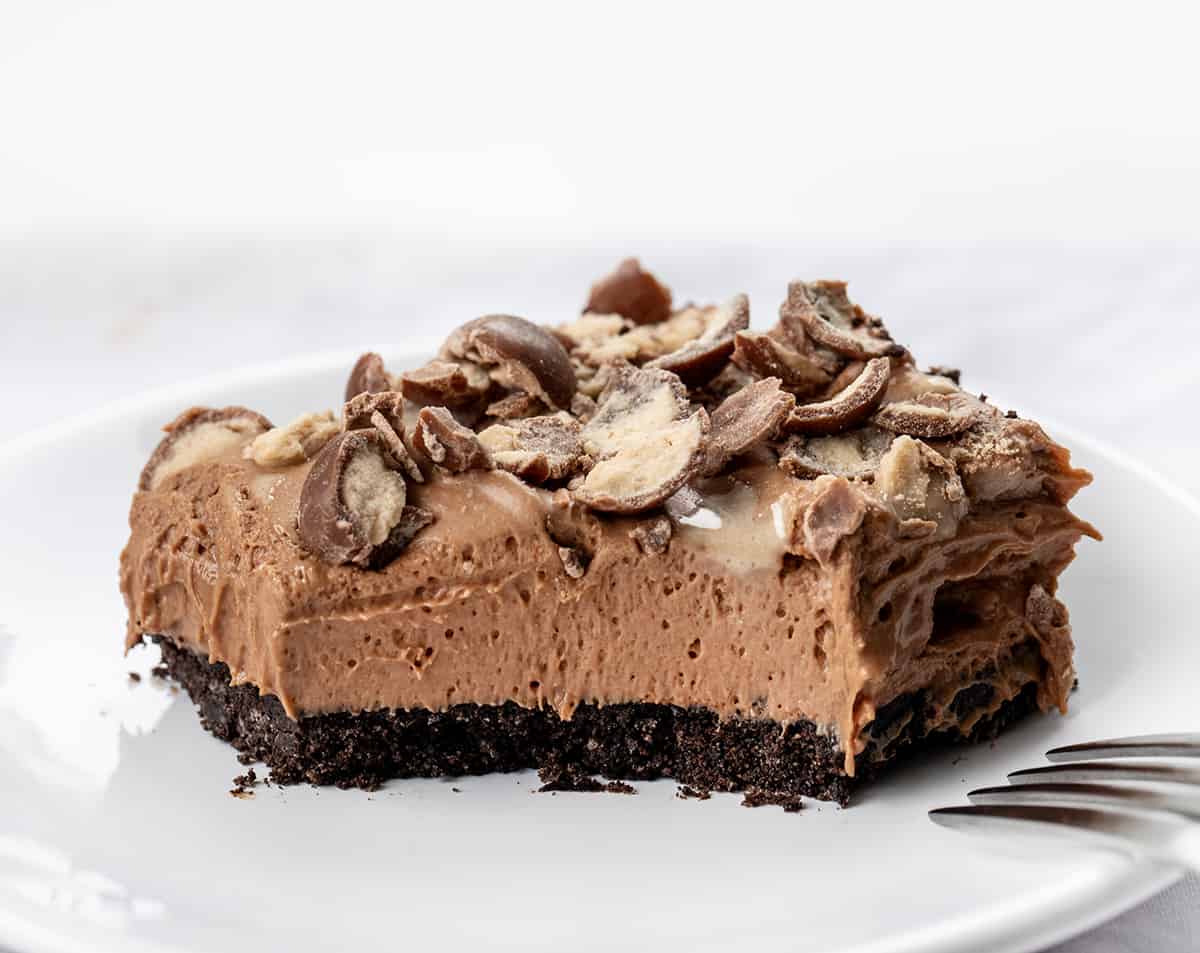 Chocolate Malt Shoppe Bars are creamy chocolatey bars with added chocolate malt powder over an Oreo cookie crust and topped with crushed malted milk ball (whopper) candies. This no-bake treat is like enjoying your favorite chocolate malted milkshake but in a dessert bar without any ice cream. Let each bite take you back to the nostalgic charm of your favorite malt shop! One Piece of Chocolate Malt Shoppe Bar on a Plate with a Bite Removed.
