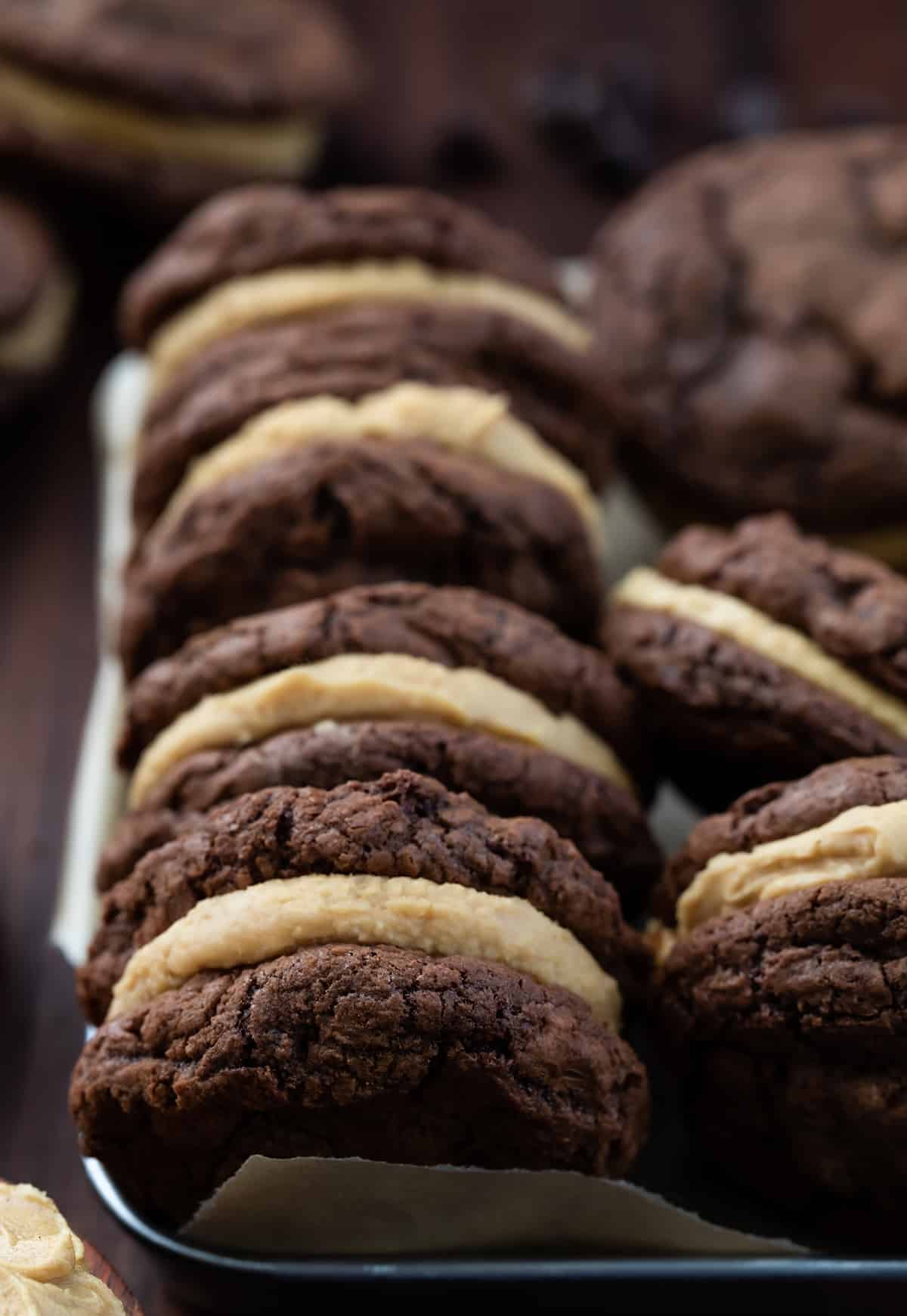 Chocolate Peanut Butter Sandwich Cookies are rich, chocolatey cookies filled with a homemade peanut butter frosting filling. The chocolate cookies are decadent enough to stand on their own with their crispy outside and soft, chewy center. (Trust me, I had a few!) But the added peanut butter filling was the perfect complement to the chocolate, making this one of my favorite sandwich cookies of all time. Rows of Chocolate Peanut Butter Sandwich Cookies in a Dish on a Dark Cutting Board.