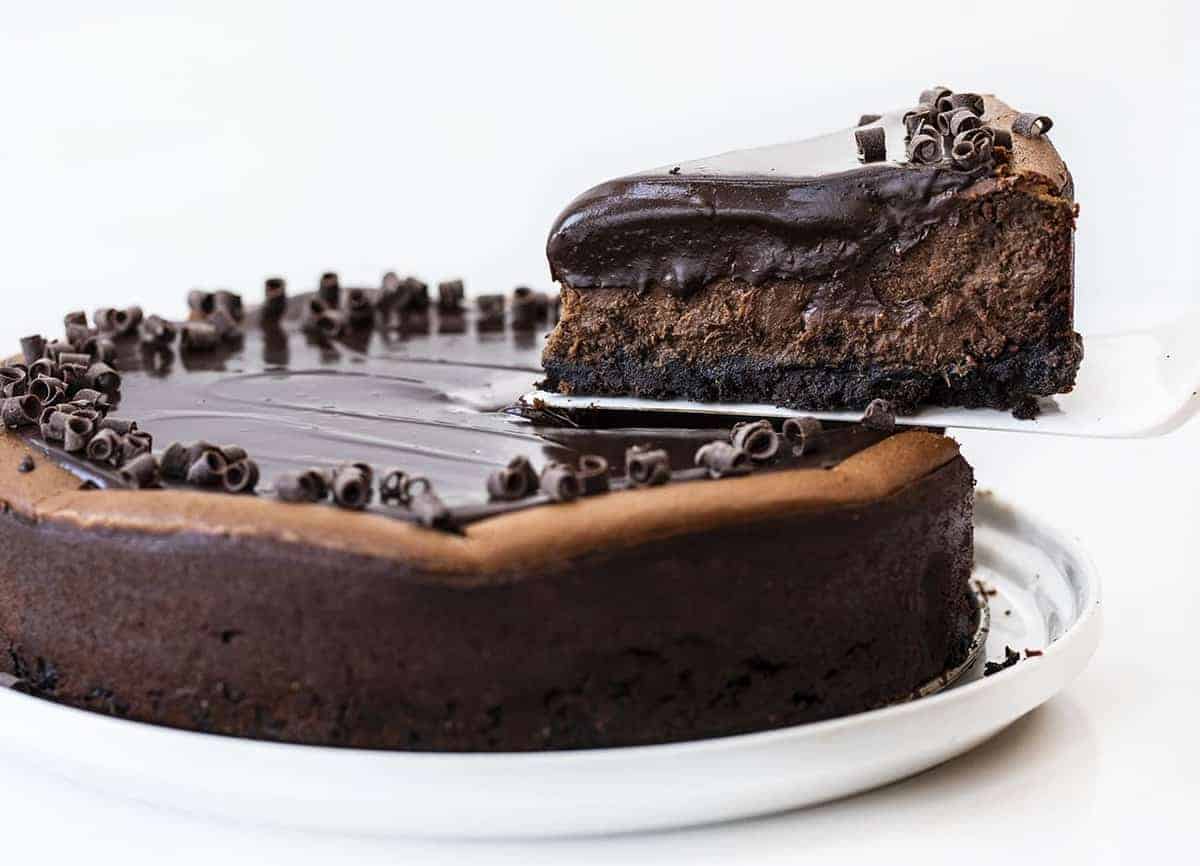 Chocolate Cheesecake is a chocolate lover's delight with an Oreo cookie crust filled in and baked with a rich and creamy chocolate cheesecake and topped with a chocolate ganache! Chocolate Cheesecake, Double Chocolate Cheesecake, Triple Chocolate Cheesecake, Ultimate Chocolate Cheesecake, Creamy Chocolate Cheesecake, How to Make Oreo Crust, How to Make Chocolate Cheesecake, baking, cheesecakes, i am baker, iambaker, Recipes One Slice of Chocolate Cheesecake Being Removed from the Pan with White Spatula